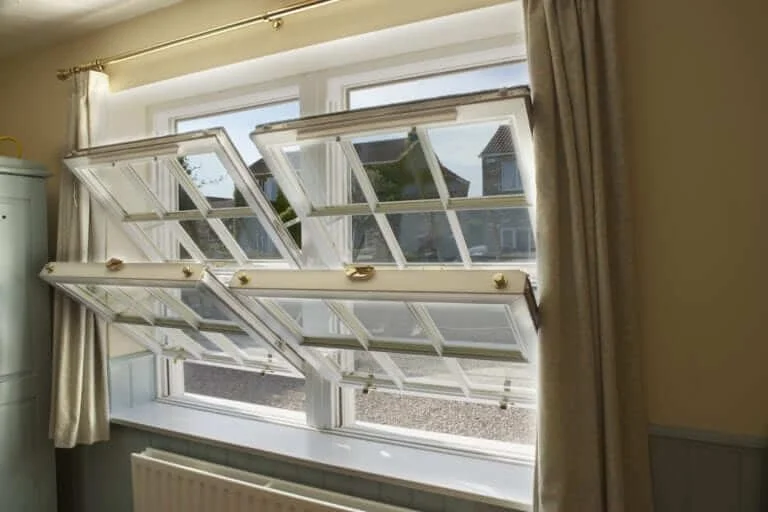 A window with three sections open outward in a residential room, letting in natural light with curtains on either side, and a view of neighboring buildings outside.