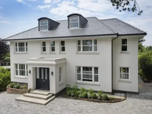 White two-story house with black front door, multiple windows, and dormer windows on the roof, surrounded by a paved driveway and landscaped bushes.