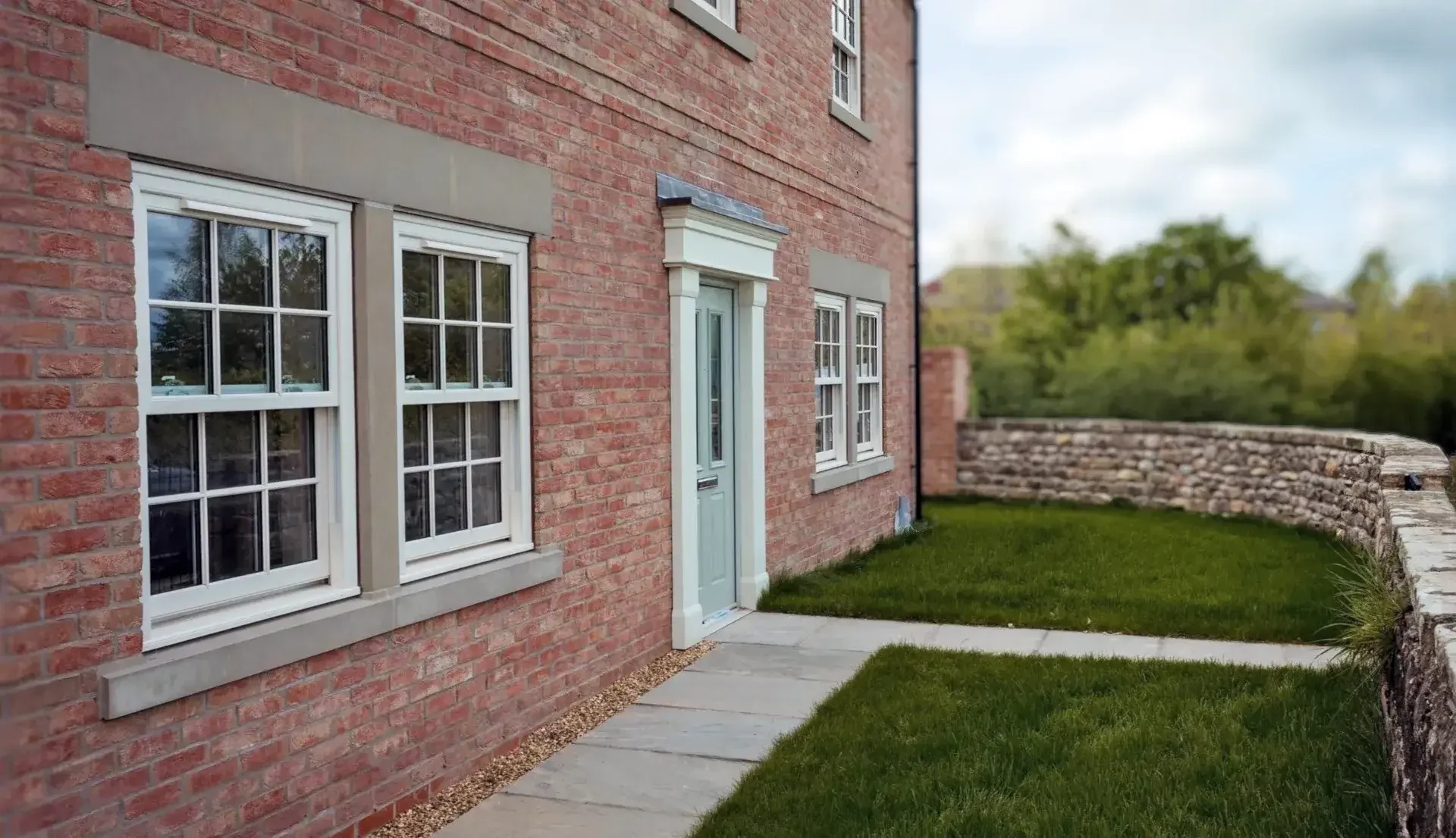 Exterior view of a red brick house with white-framed windows, a mint green door, and a stone pathway leading to the door. There is a green lawn and a stone wall in the background.