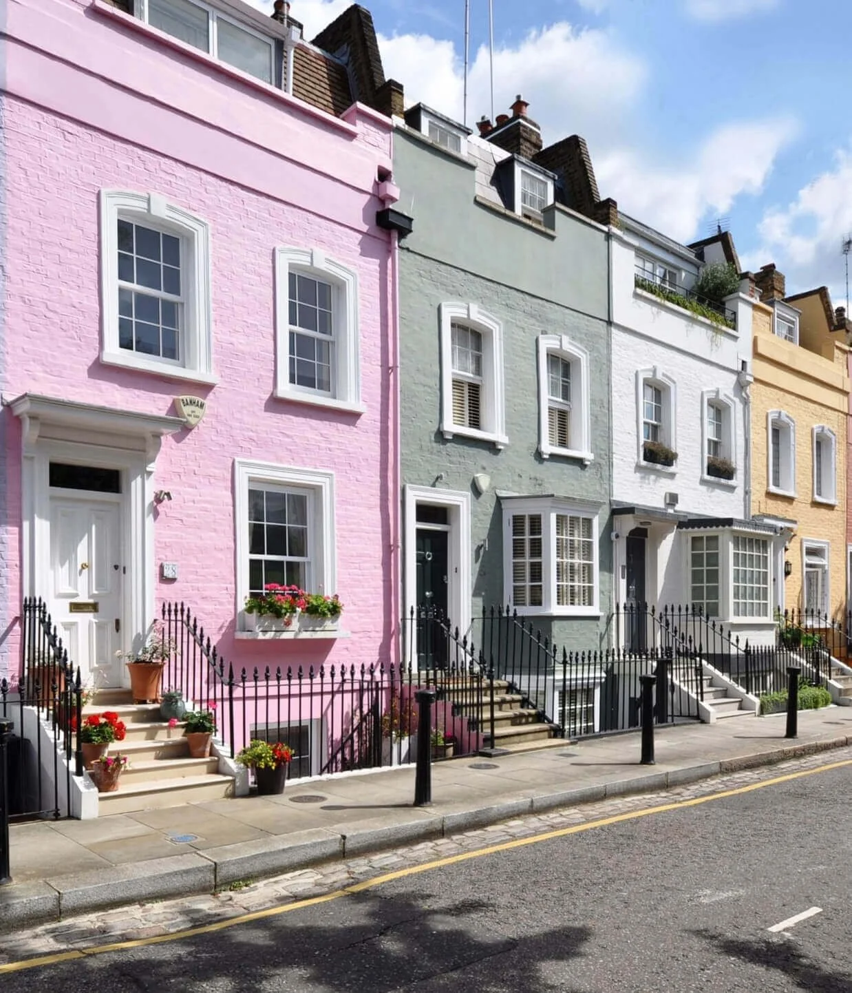 Colorful row of houses with pastel pink, green, white, and yellow facades, each with steps, black railings, and potted flowers at the entrances, under a partly cloudy sky.
