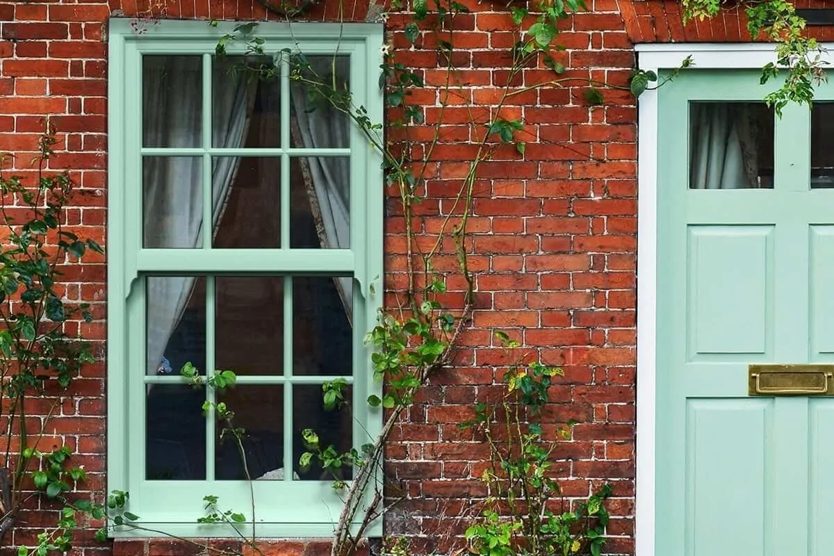 A red brick house exterior featuring a light green window with divided panes and a matching green door with a mail slot, surrounded by climbing green vines.