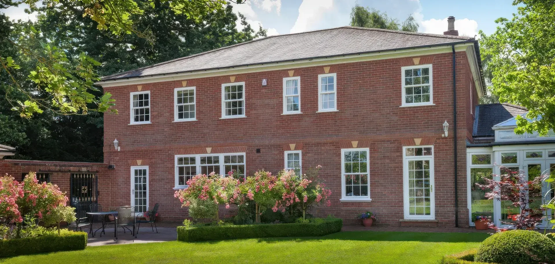 A two-story brick house with large windows, a sloped roof, and a garden with pink flowering bushes and green grass.