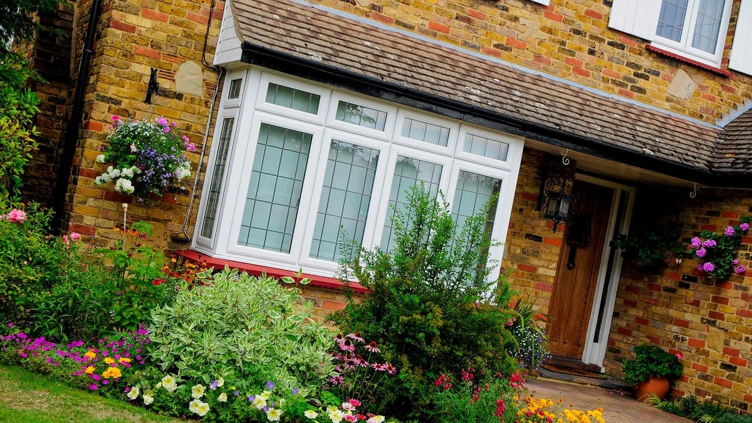 A brick house with a tree and colorful flowers in front of the entrance and a bay window.