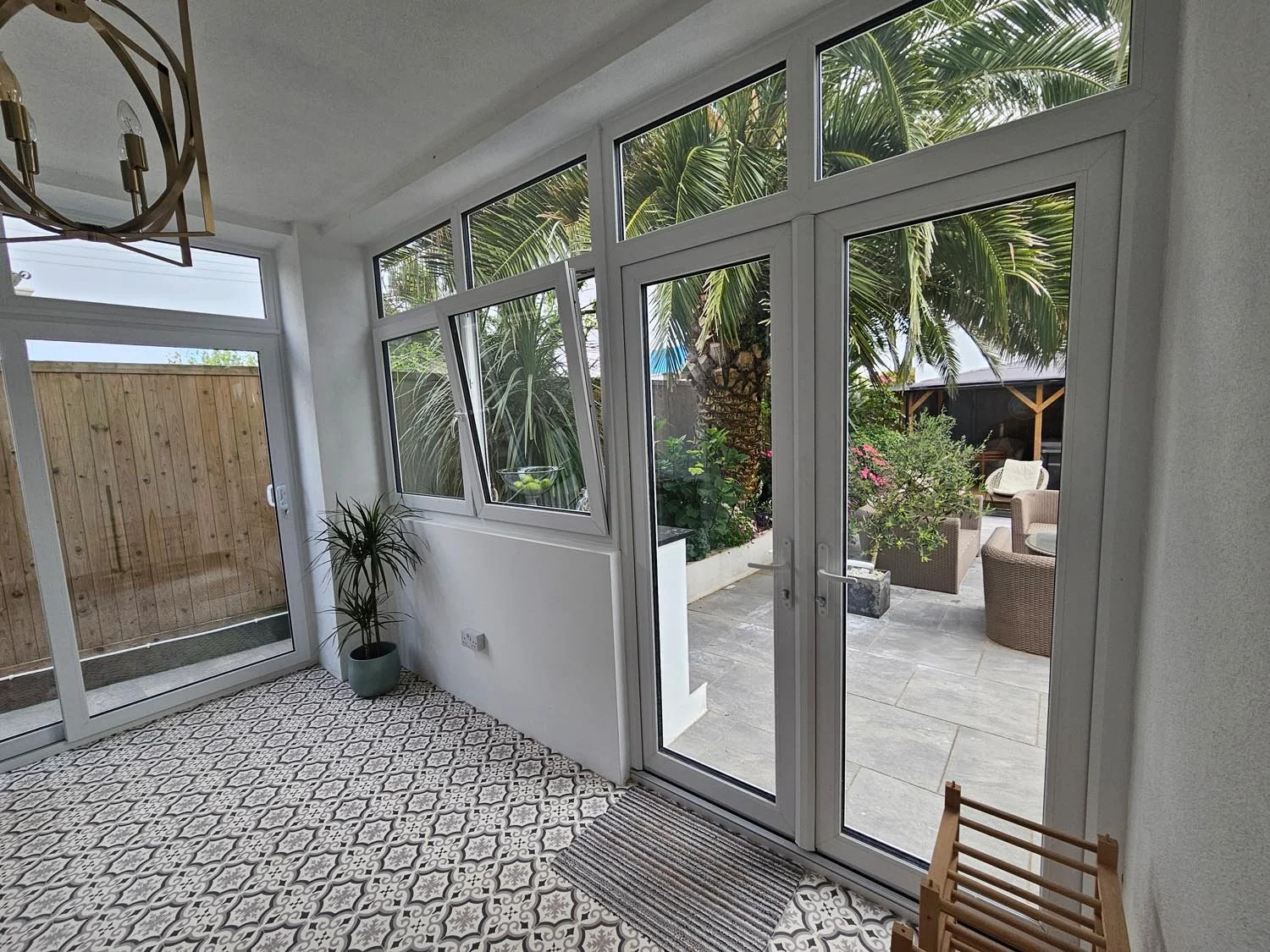View of a sunroom with patterned tile flooring, white walls, and large glass doors leading to a backyard with garden furniture and a tall palm tree.