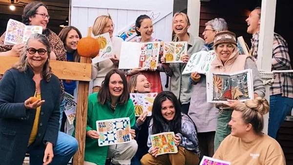 A joyful group of artists standing on a barn porch, laughing and holding up colorful sketchbook paintings — sharing about The Fellow Artists Group, a supportive creative community on Patreon.