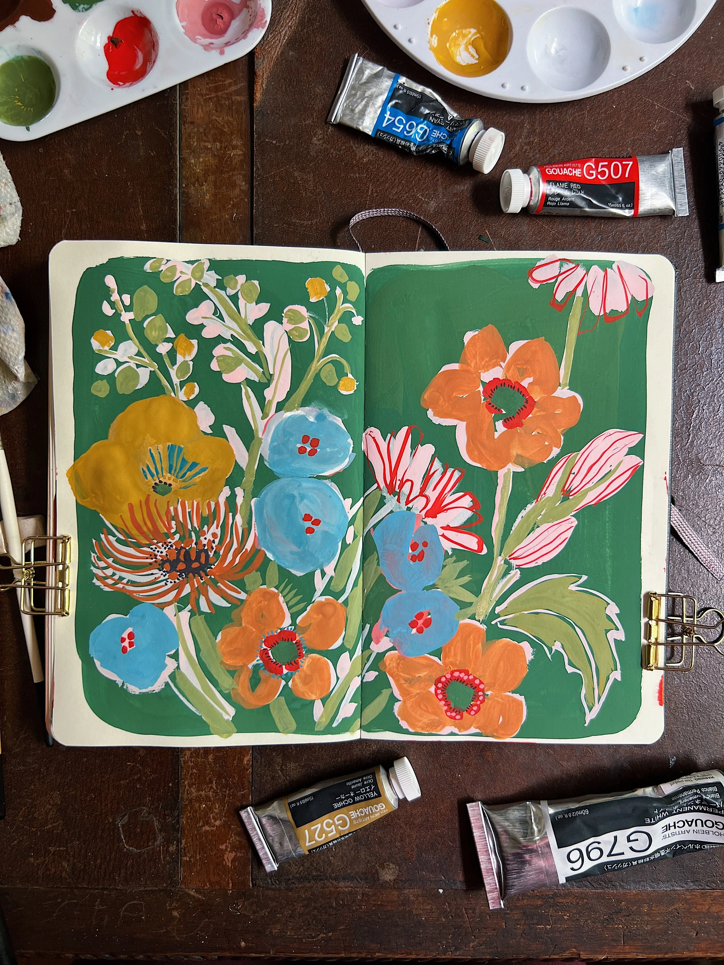 An open sketchbook shows colorful and folksy flowers painted in gouache