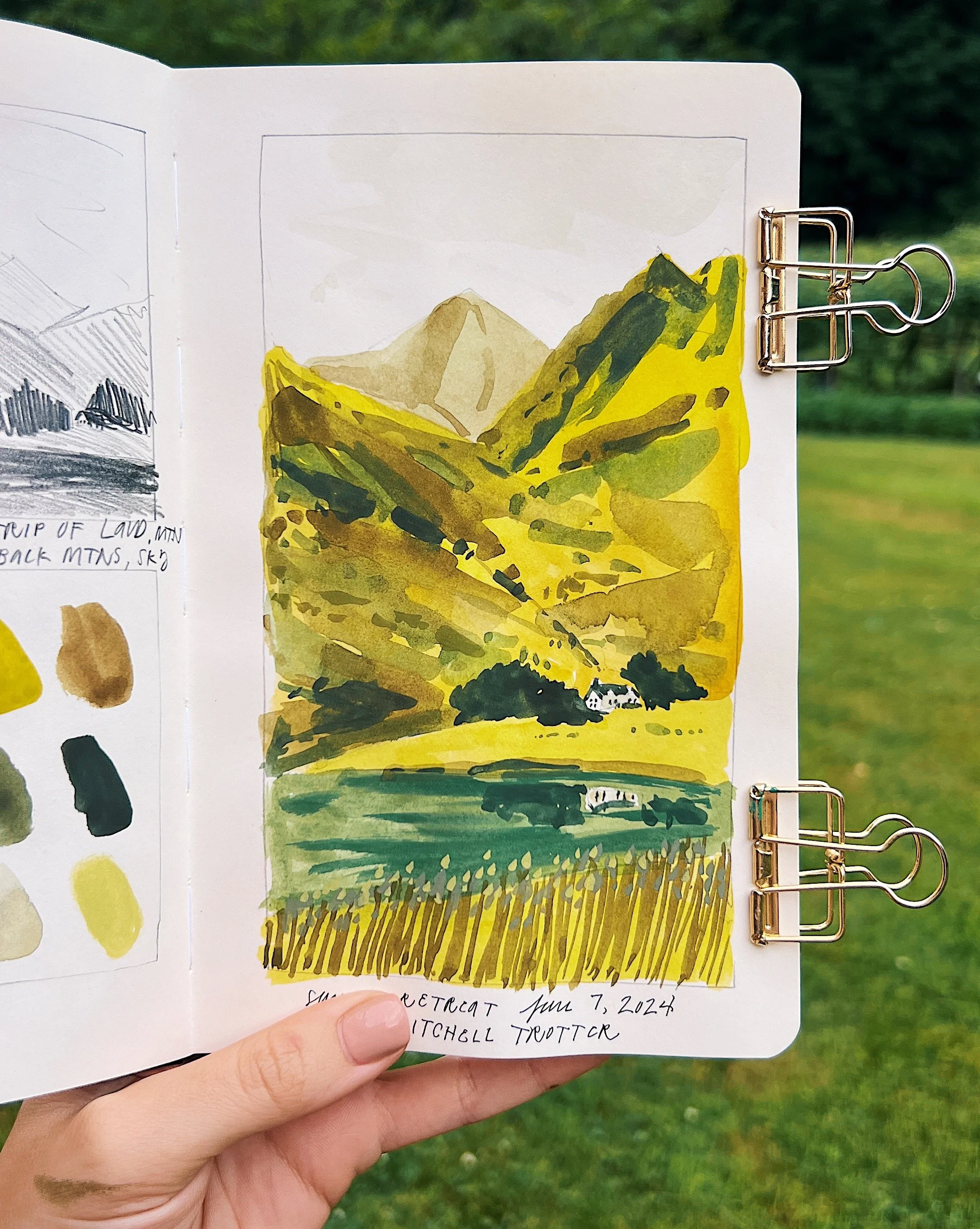 Dylan's hand holds up her sketchbook showing a grassy and mountainous landscape painting done in gouache, featuring a tiny white house.