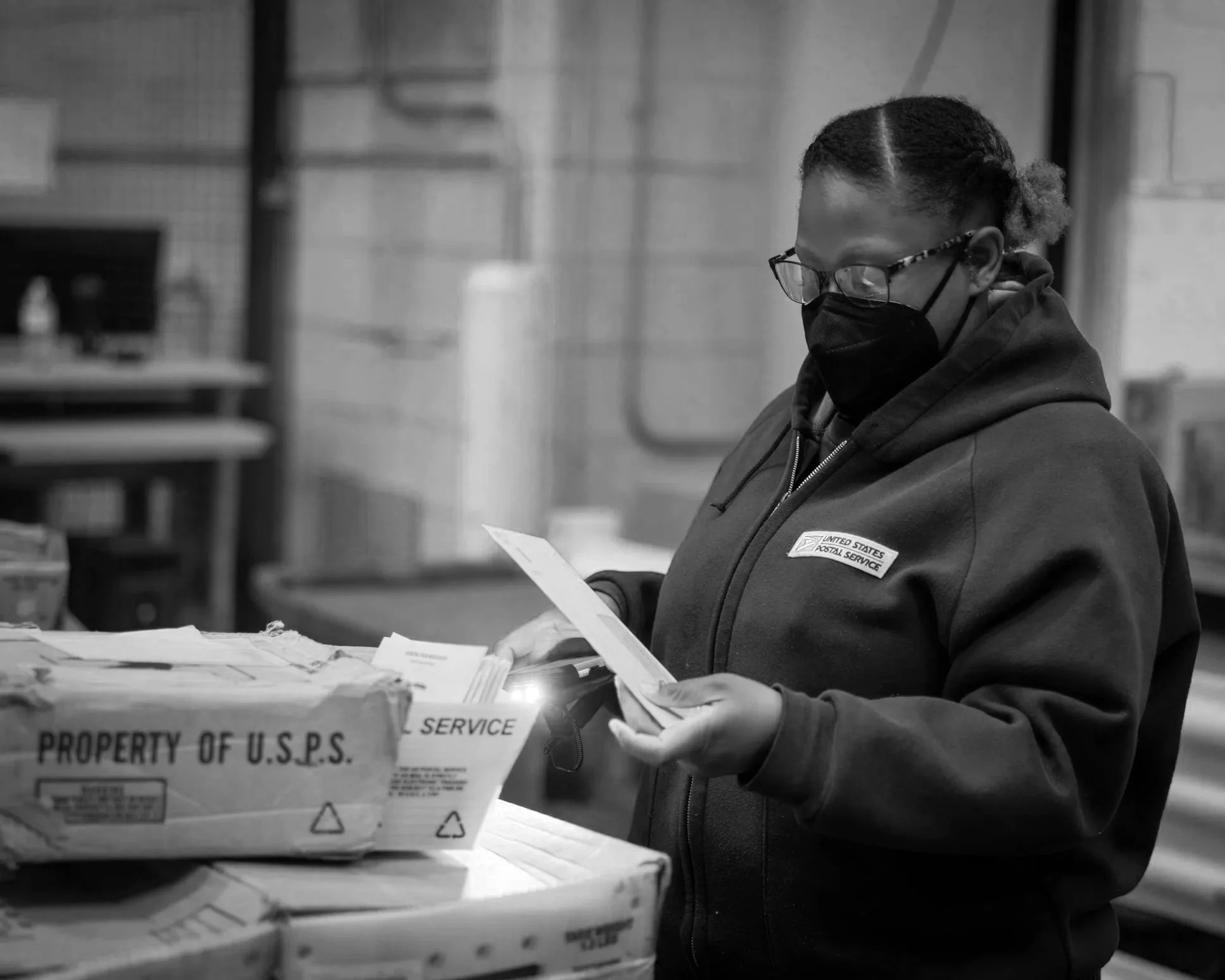 A person wearing glasses and a black mask, dressed in a United States Postal Service jacket, is processing mail or packages in a postal warehouse.