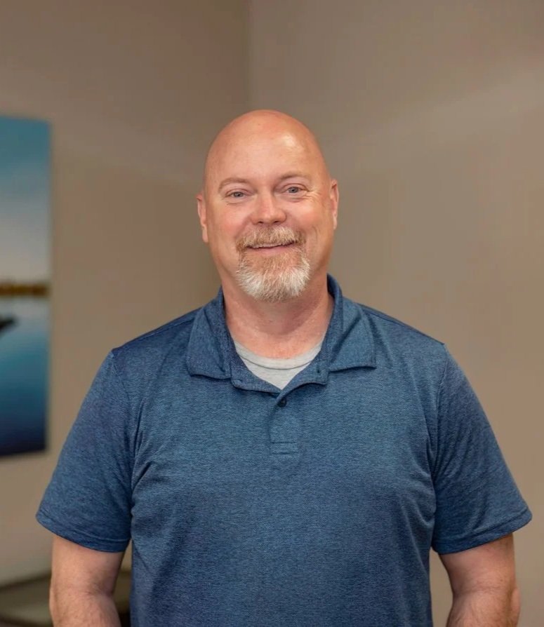 A middle-aged man with a shaved head, goatee, and blue eyes, wearing a blue collared shirt, smiling indoors against a beige wall with a framed nautical photo in the background.