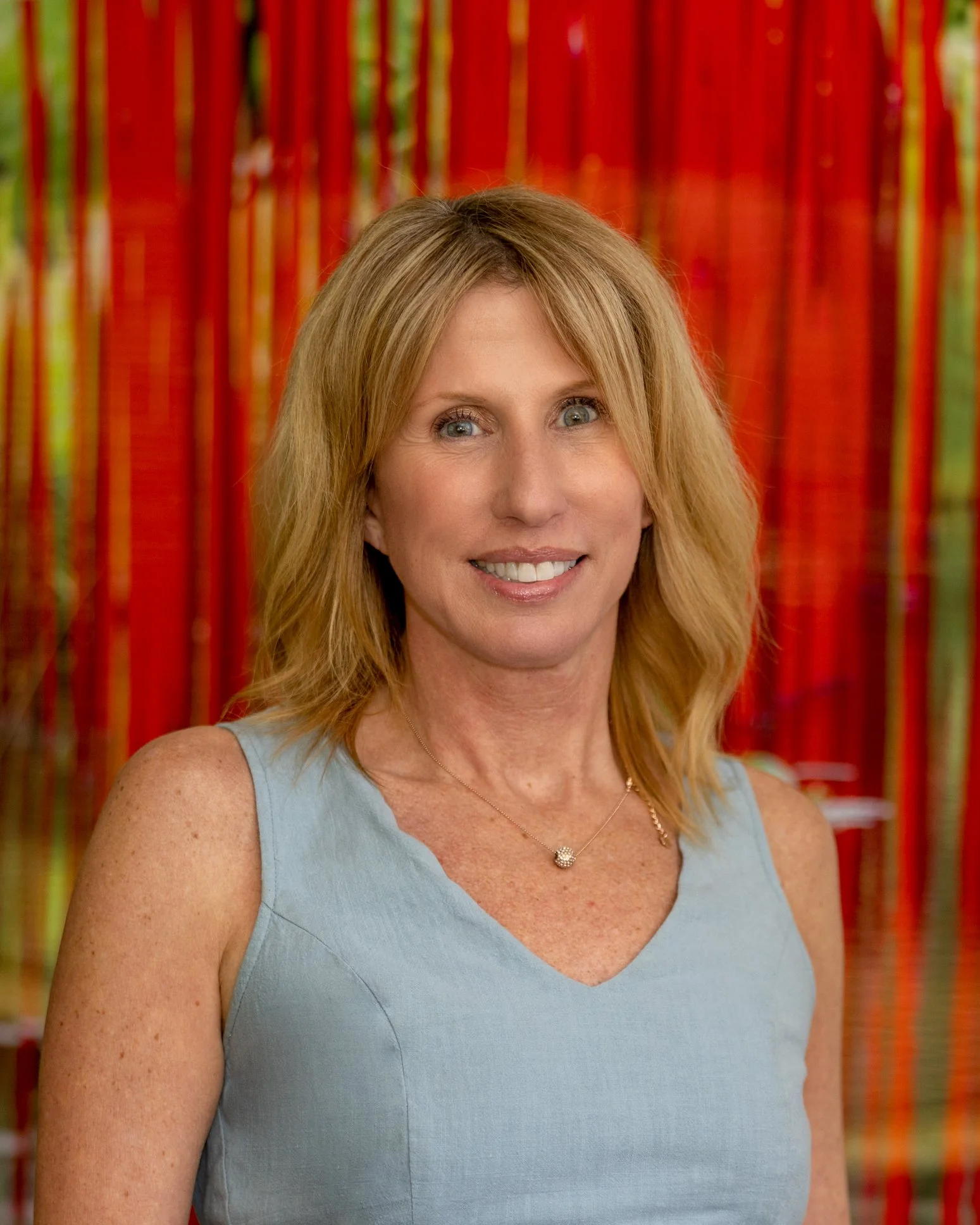 A woman with blonde hair, wearing a light blue sleeveless top and a necklace, smiling at the camera, with water reflecting trees in the background.