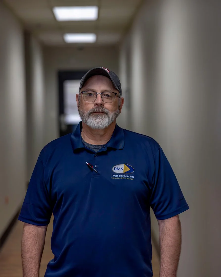 A middle-aged man with glasses and a beard wearing a navy blue polo shirt with an embroidered logo that says DMS, standing in a hallway with beige walls and rectangular ceiling lights.