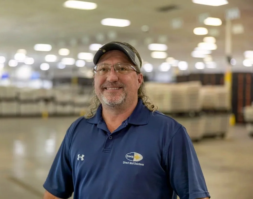 A man with glasses, wearing a black visor and a blue polo shirt with a logo, standing inside an industrial warehouse or mail sorting facility.