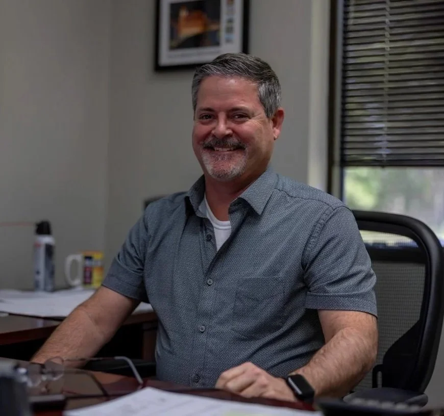 A middle-aged man with salt-and-pepper hair and a beard, smiling and seated at a desk in an office. He is wearing a short-sleeved, button-up gray shirt and a smartwatch on his left wrist. The office has a window with blinds, a framed picture on the wall, and various objects on the desk, including papers, a mug, and personal care items.