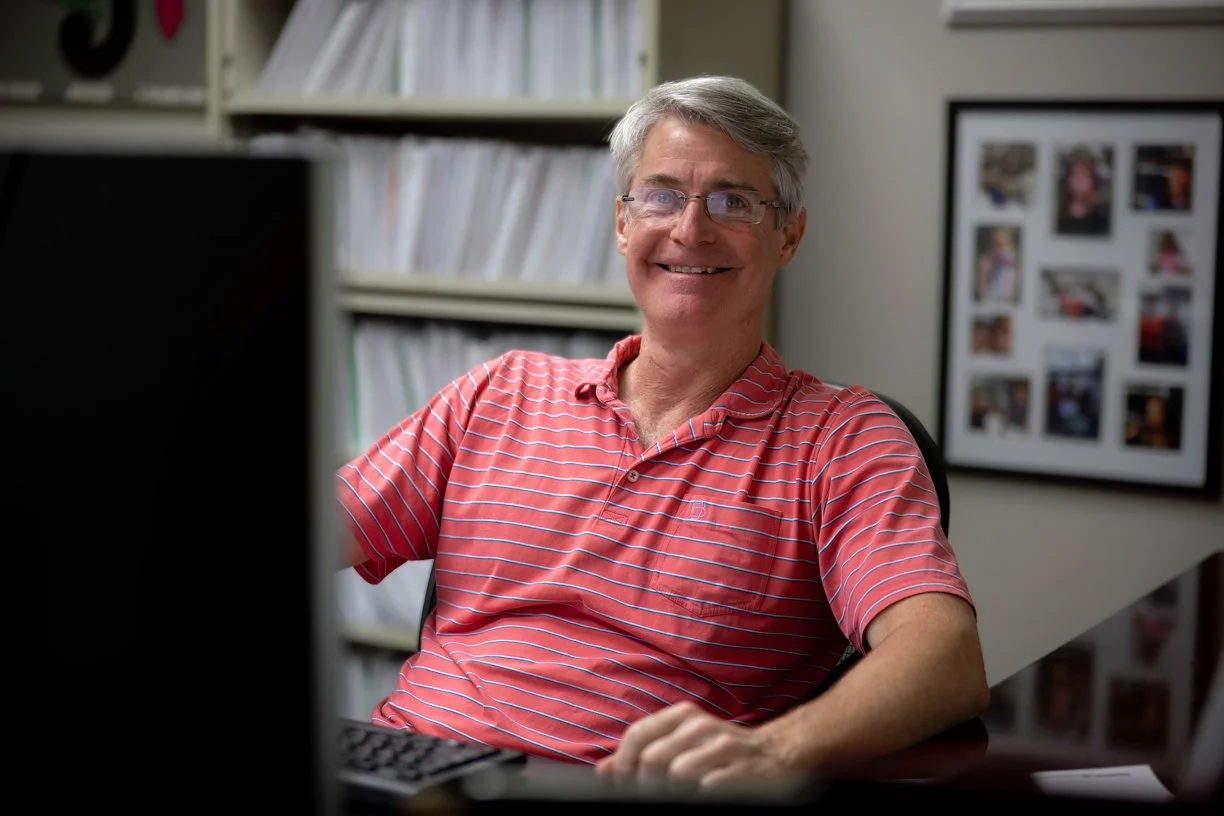 Smiling middle-aged man wearing glasses and a red striped polo shirt sitting at a desk in an office, with shelves of files behind him and a framed photo collage on the wall.
