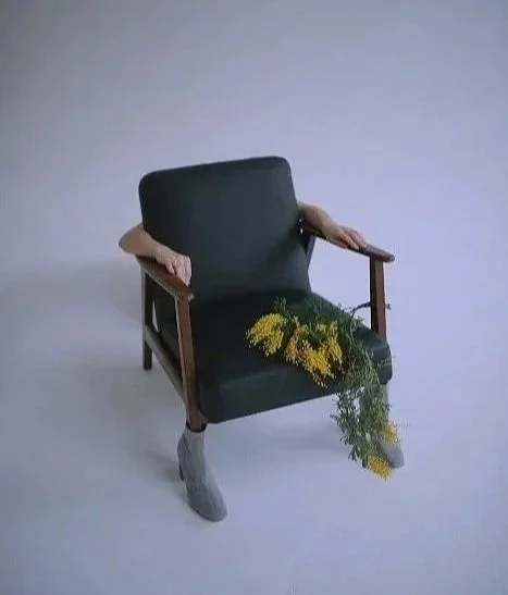 An old armchair with human arms and legs, with flowers hanging from the chair's seat.