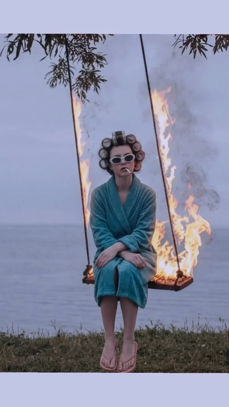 A woman sitting on a swing near the water, with a fire burning behind her on the swing, wearing sunglasses, curlers in her hair, a blue robe, and sandals, holding a cigarette.