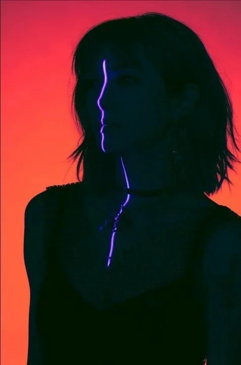 A person with shoulder-length dark hair standing against a colorful sunset background, illuminated by purple light streaks on their face and neck.
