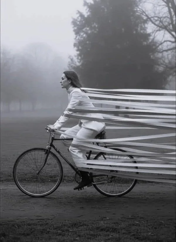 A woman in a white suit is riding a bicycle in an outdoor park with leafless trees and foggy weather, and her body appears to be wrapped in or surrounded by streaming white ribbons.
