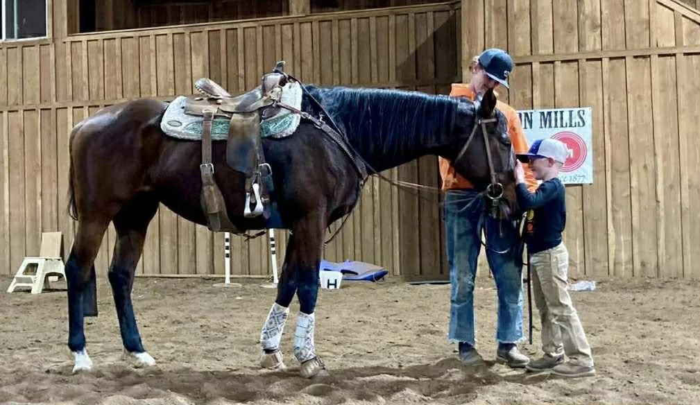A person wearing an orange shirt and blue hat assisting a young boy with a gray cap in holding a brown horse with a saddle in an indoor riding arena with wooden walls.