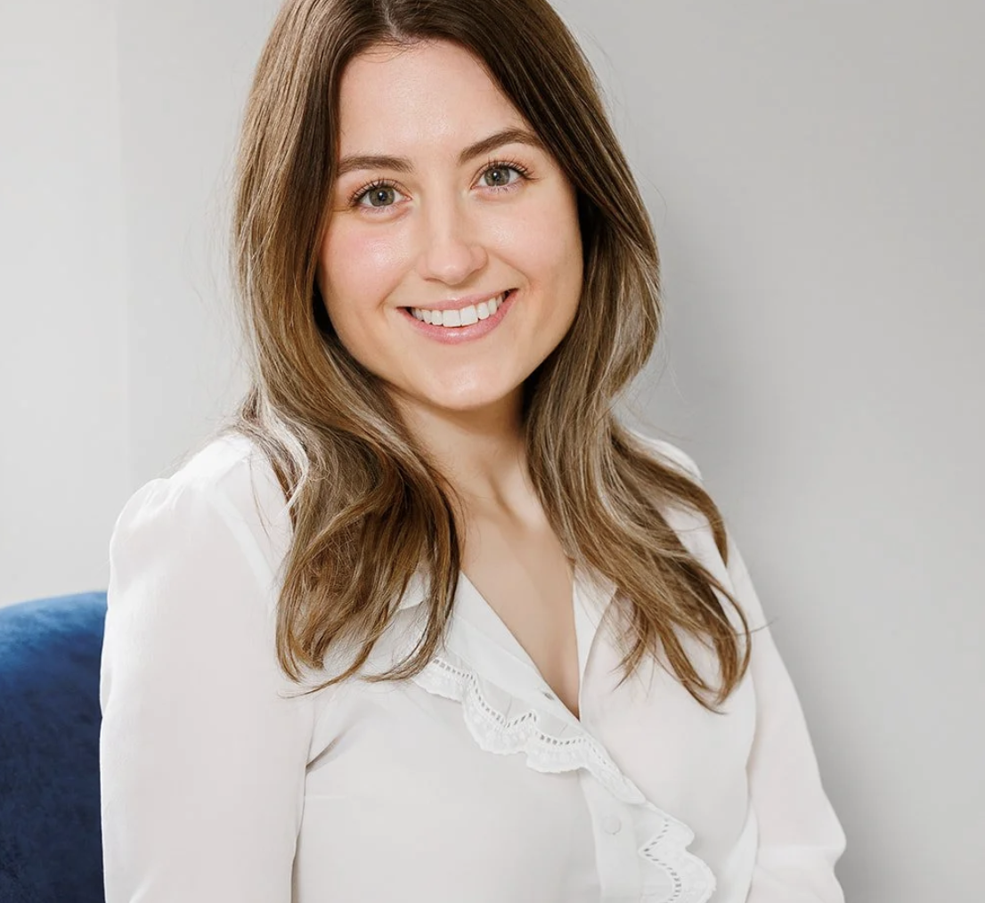 A young woman with long brown hair and a white blouse smiling at the camera.