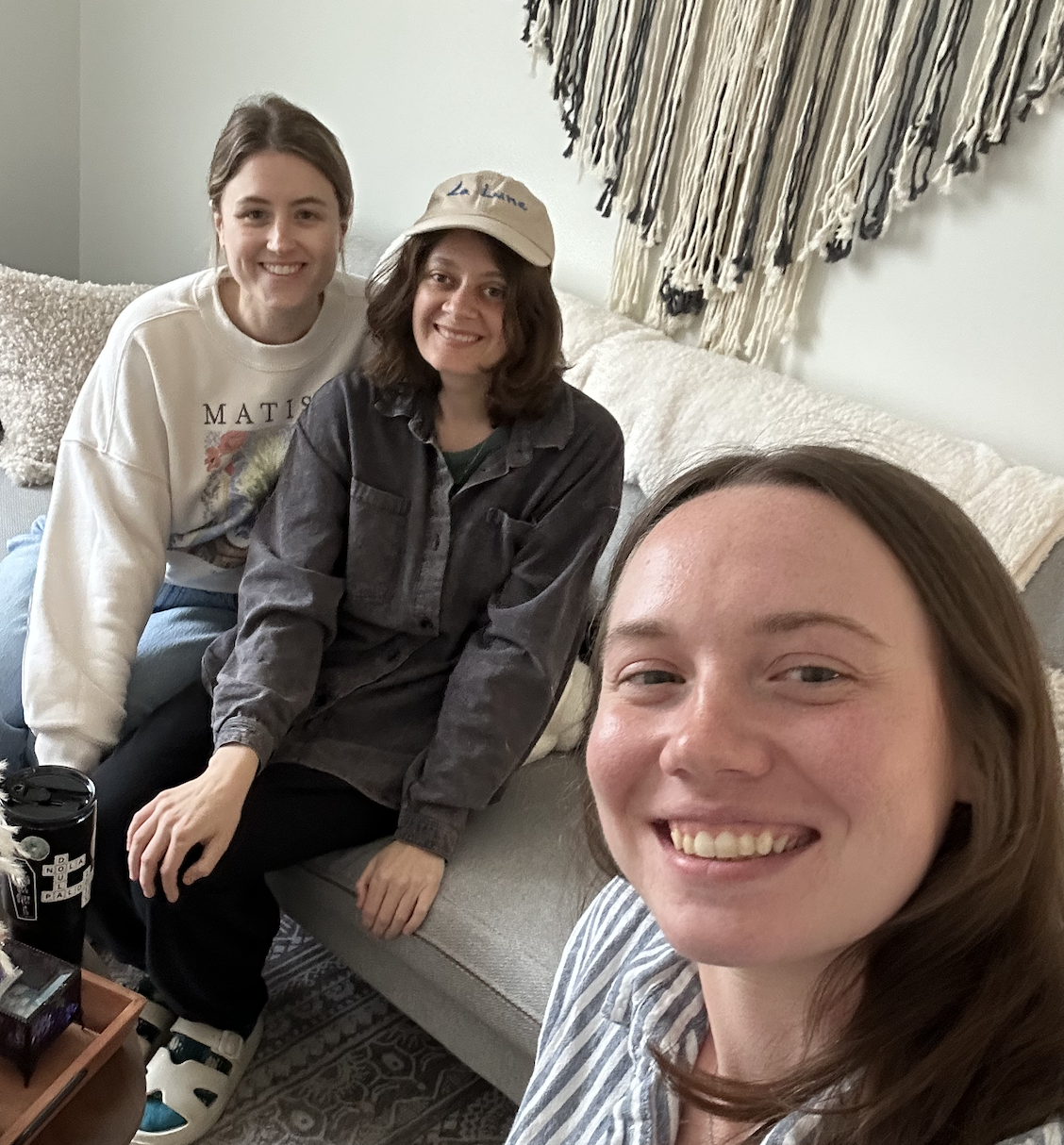 Three women smiling and taking a selfie in a cozy living room with a beige couch, a textured throw blanket, and a decorative wall hanging.