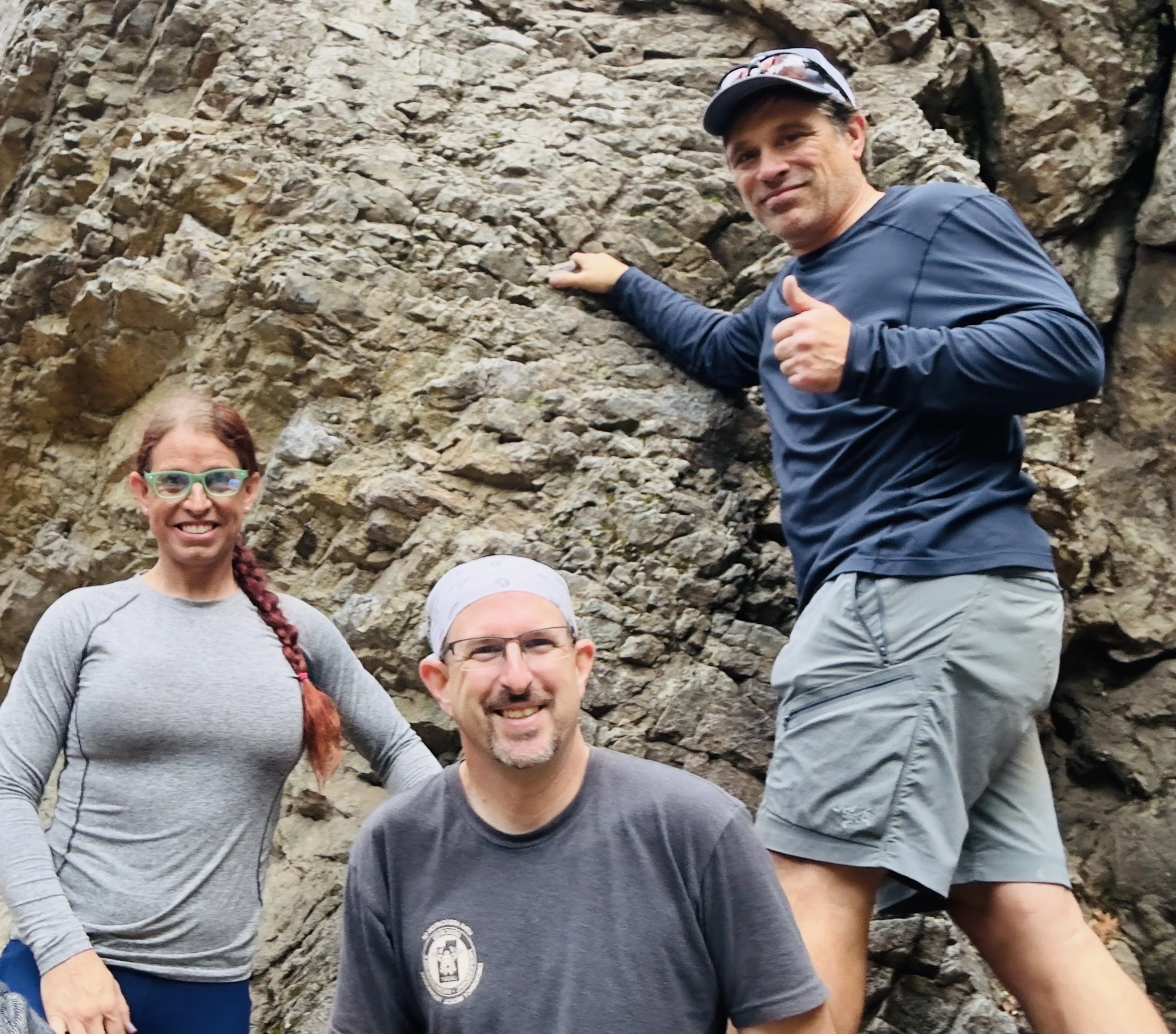 Three people posing outdoors in front of a rocky cliff, smiling and dressed in casual hiking or outdoor clothing.
