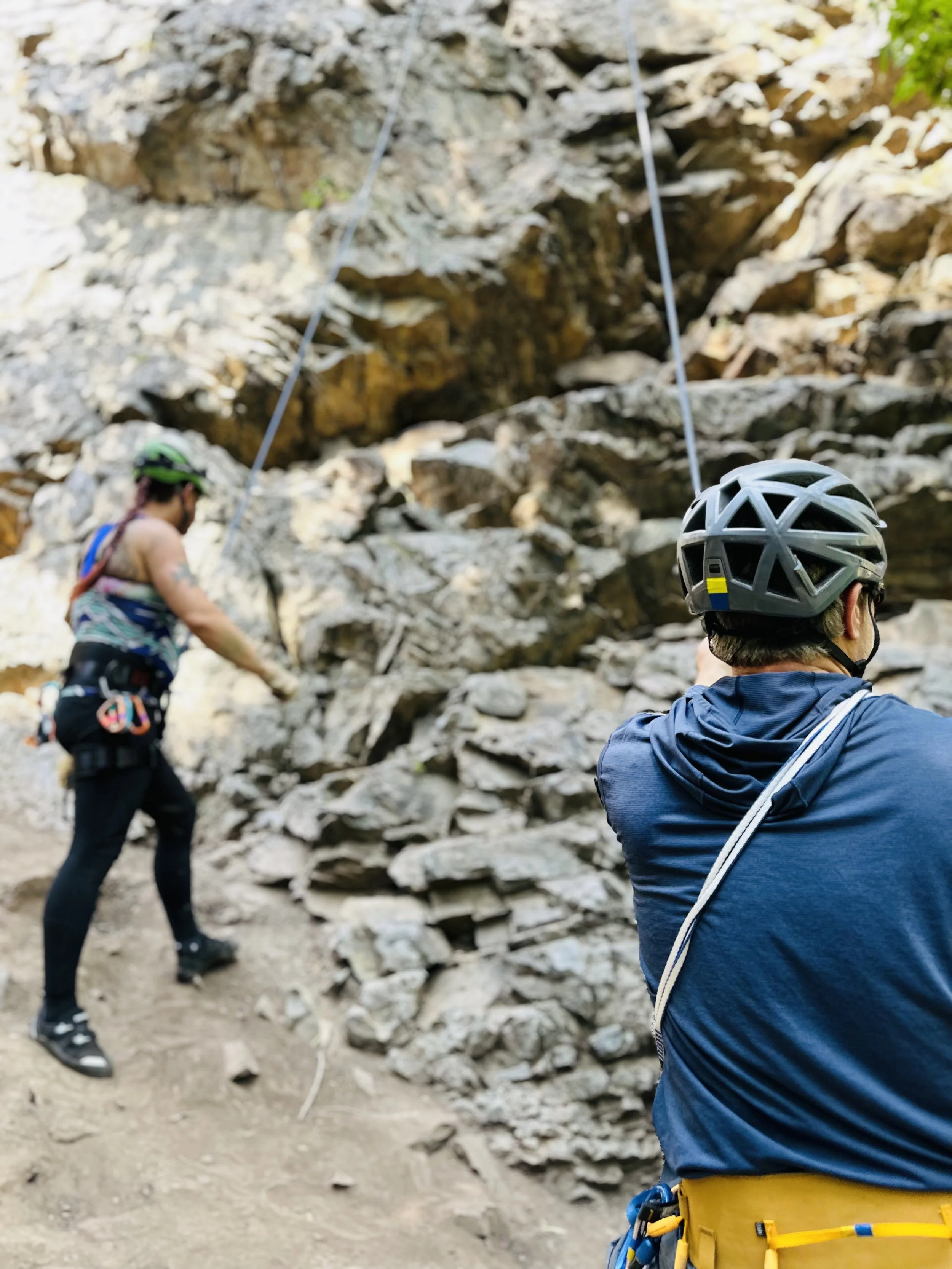 Two rock climbers wearing helmets and harnesses, preparing to climb a rocky outdoor wall.