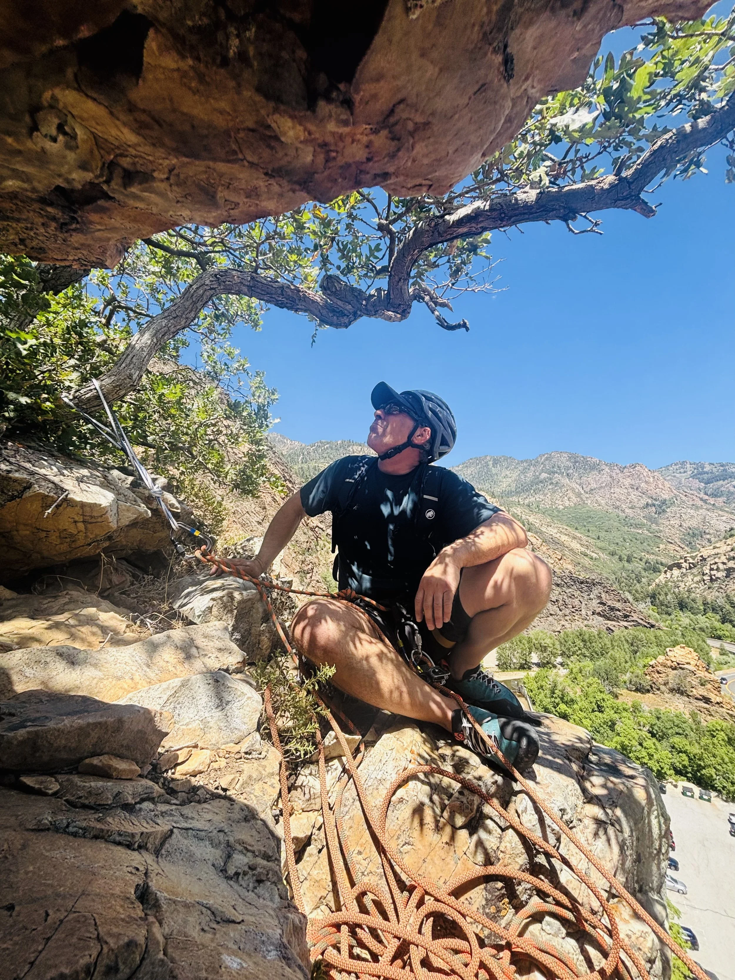 A man wearing a helmet and sunglasses climber climbing a rocky outdoor wall with mountain and blue sky in the background.