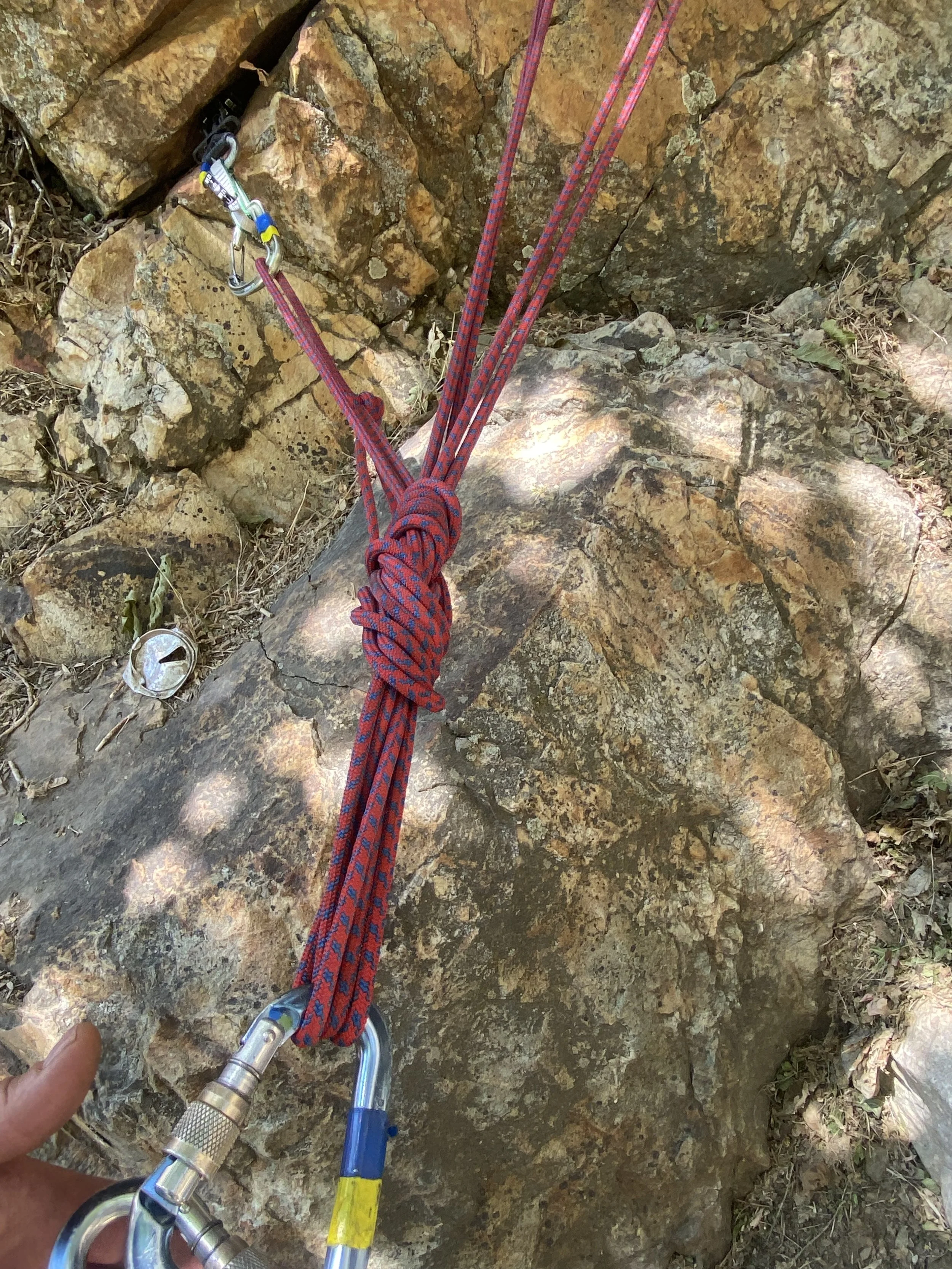 Climbing gear including a red and blue rope secured with a carabiner and a quickdraw, attached to a rock climbing anchor on a rugged rocky surface in an outdoor setting.