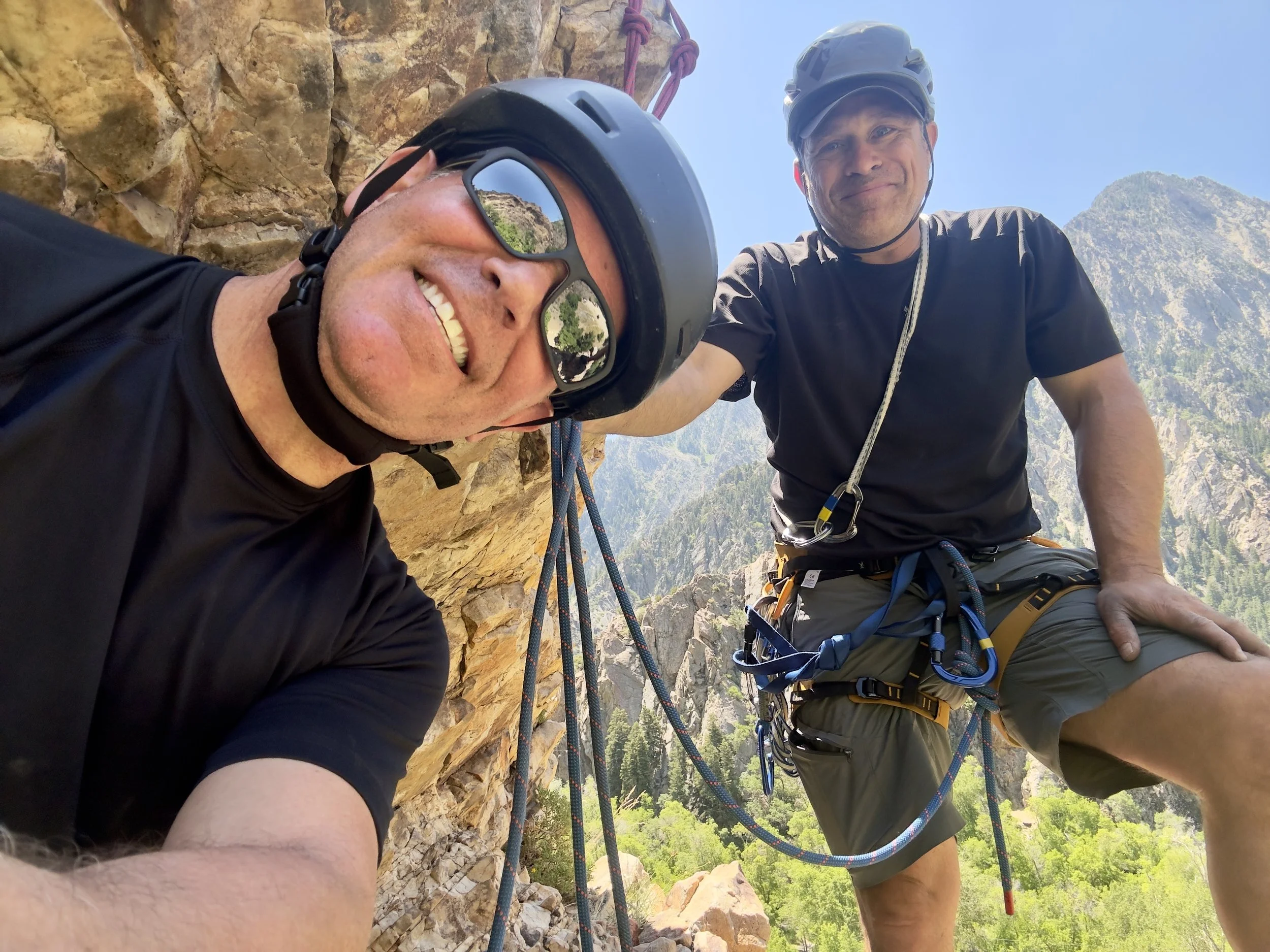 Two men in climbing helmets and gear taking a selfie during a rock climbing or mountaineering adventure with mountain landscape in the background.