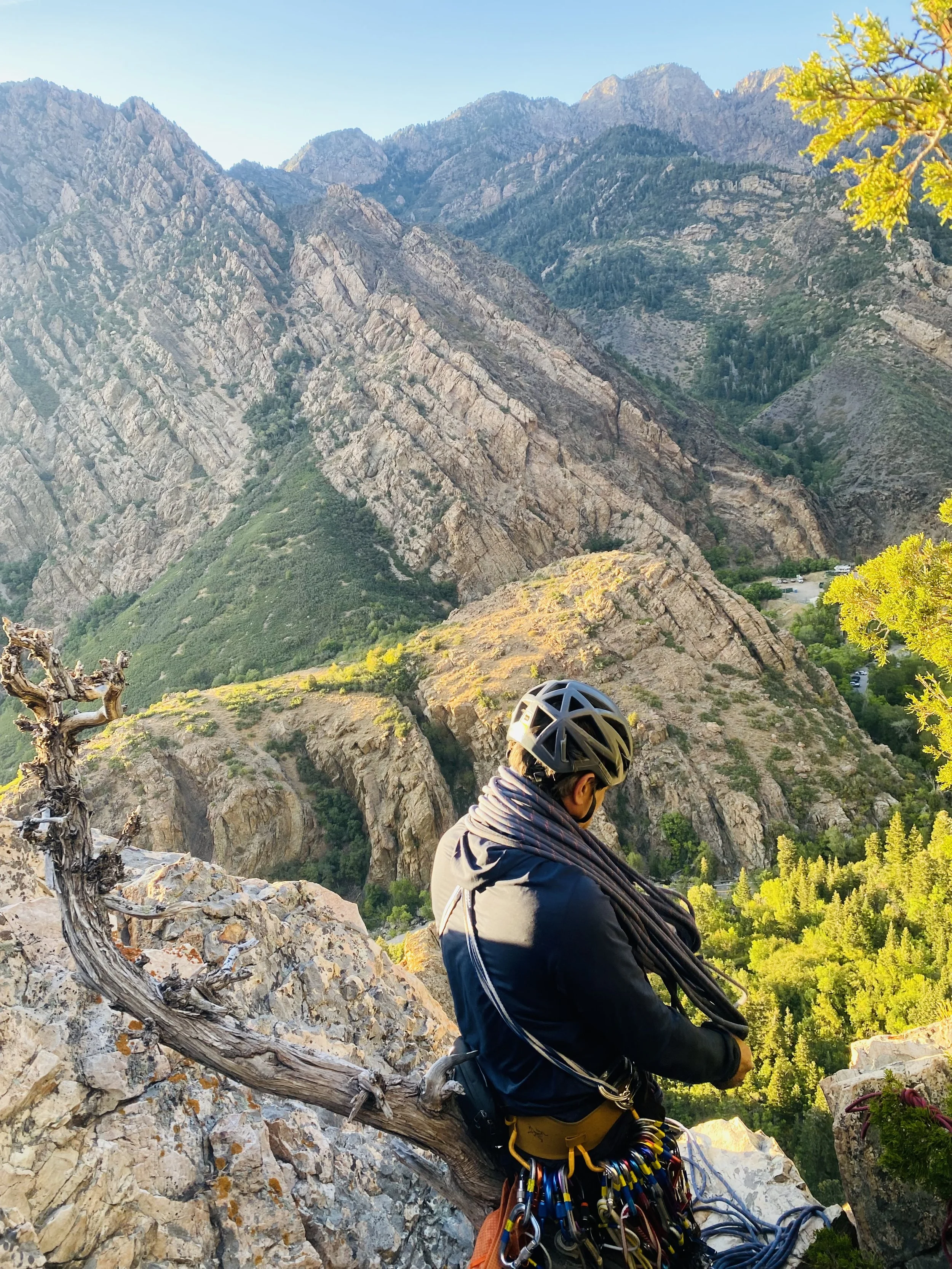 A rock climber with a helmet and climbing gear sits on a rocky ledge overlooking a valley with steep mountain slopes and green trees.
