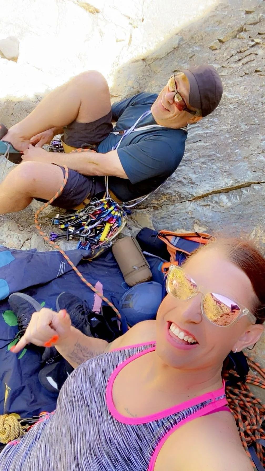Two rock climbers taking a selfie, resting against a rock wall with climbing gear around them. The woman in the foreground is smiling and wearing sunglasses, a gray athletic top with pink trim, and black pants. The man in the background is lying agai