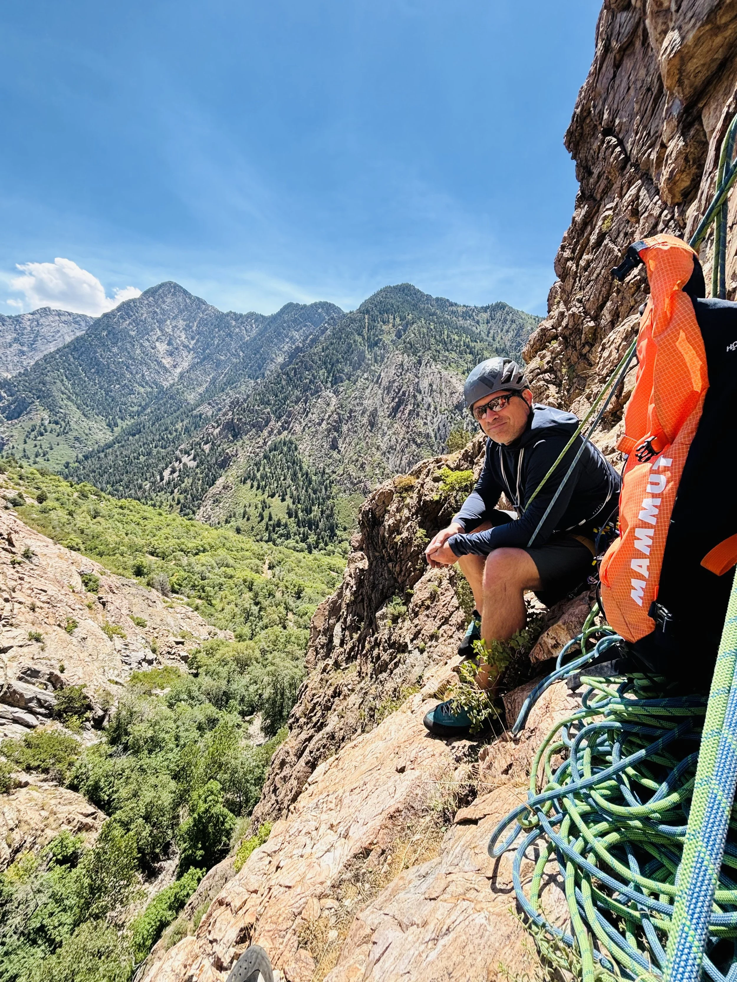 A man wearing a helmet and sunglasses taking a break on a steep mountain rock face, with mountains and green forest below, and climbing gear and a large orange backpack nearby.