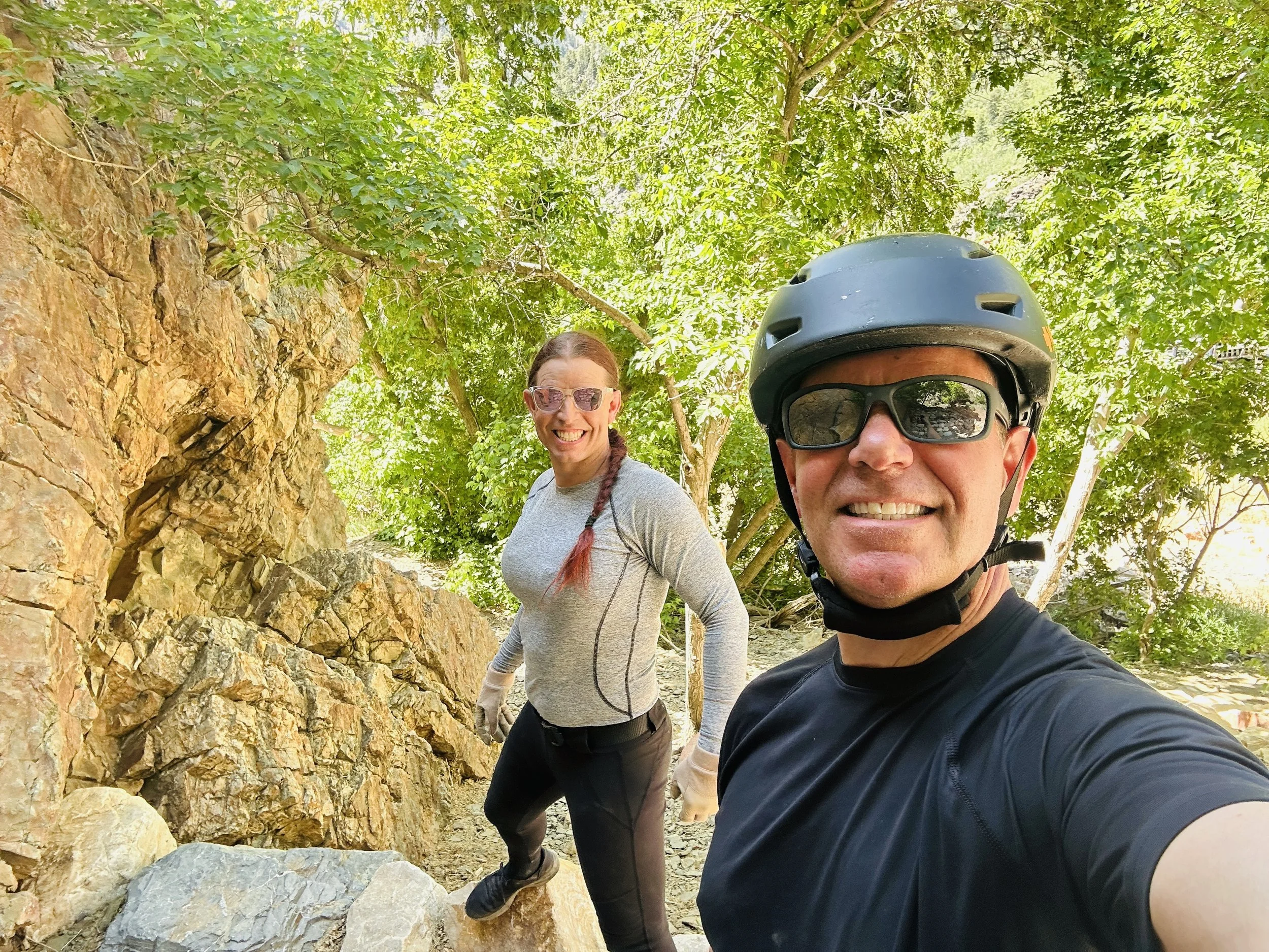 A man and woman outdoors on a rocky trail surrounded by green trees, taking a selfie. The man is wearing a helmet and sunglasses, and the woman is wearing glasses and a light-colored athletic shirt.