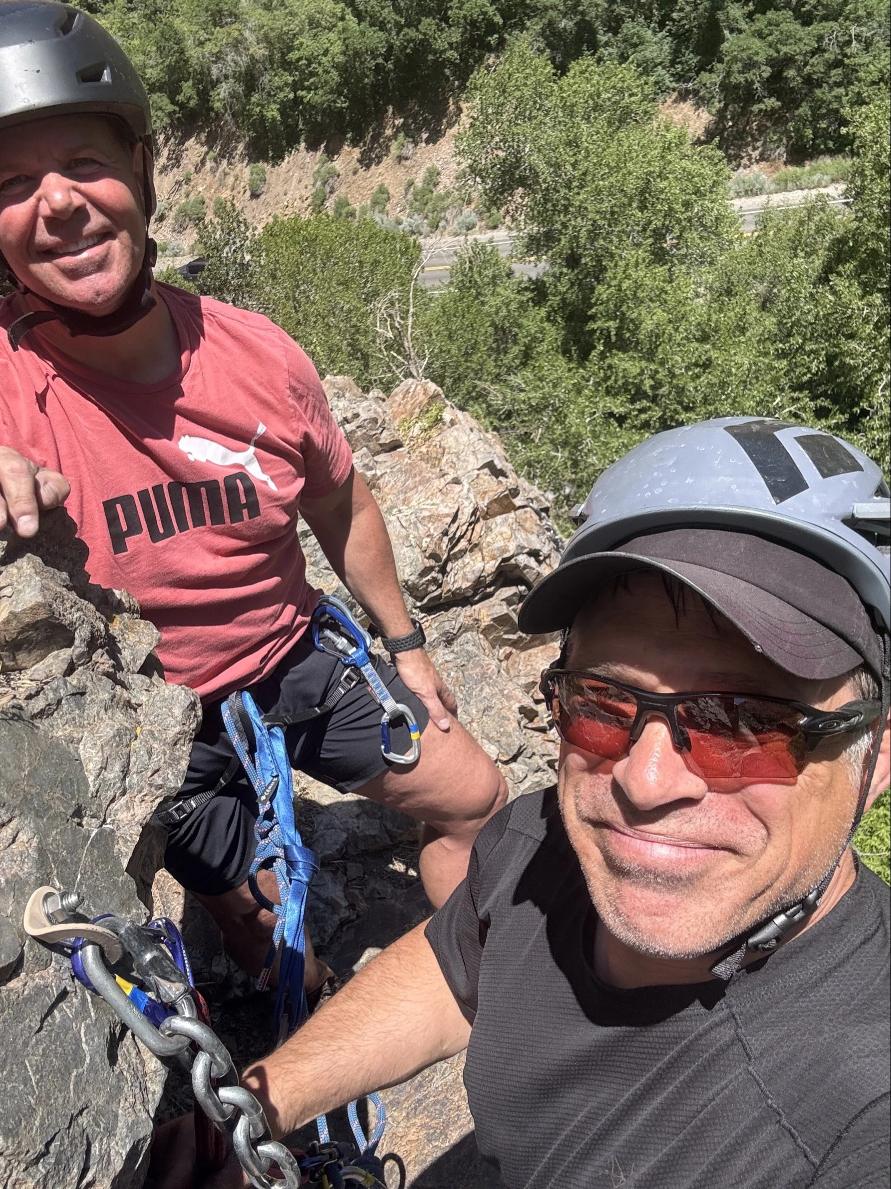 Two men in outdoor gear and helmets climbing a rocky outdoor trail surrounded by trees, with greenery and a trail visible in the background.