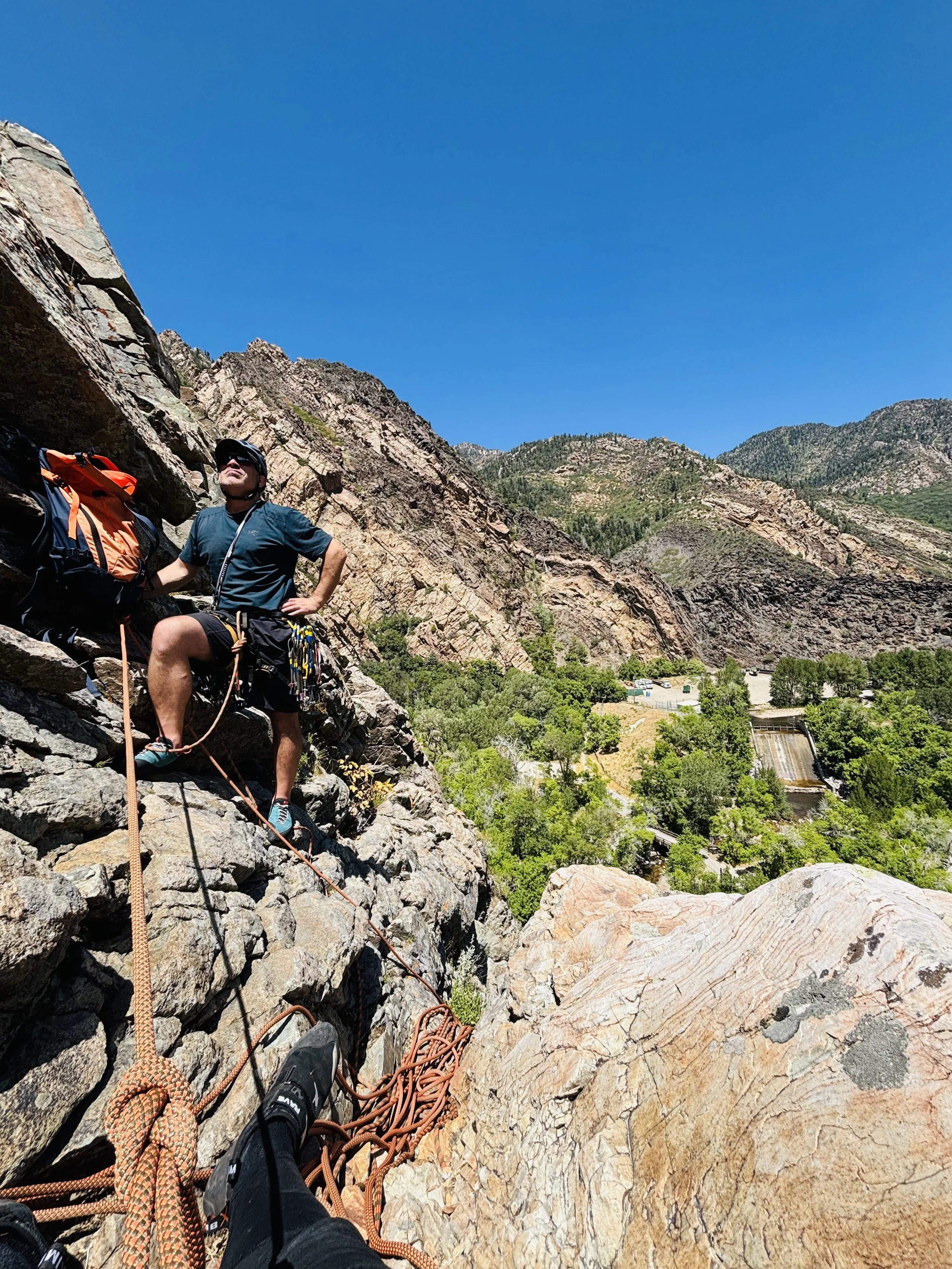A person climbing a rocky mountain with climbing gear, ropes, and a backpack on a bright sunny day, overlooking a valley with trees and a small dam or waterfall in the distance.