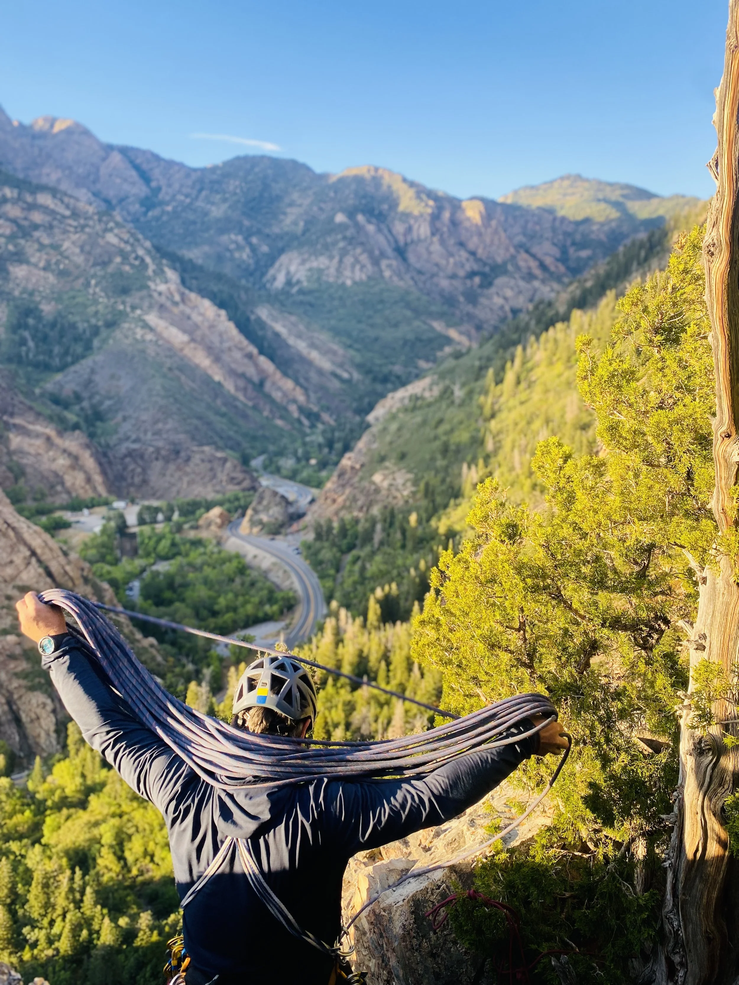 A person wearing a helmet and harness is rappelling down a rock cliffside with a scenic view of a valley, mountains, and a winding road below.