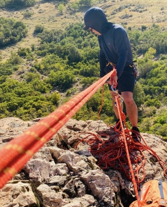 Person in black helmet and hoodie, standing on rocky terrain, engaged in rock climbing or rappelling, with ropes and safety gear.