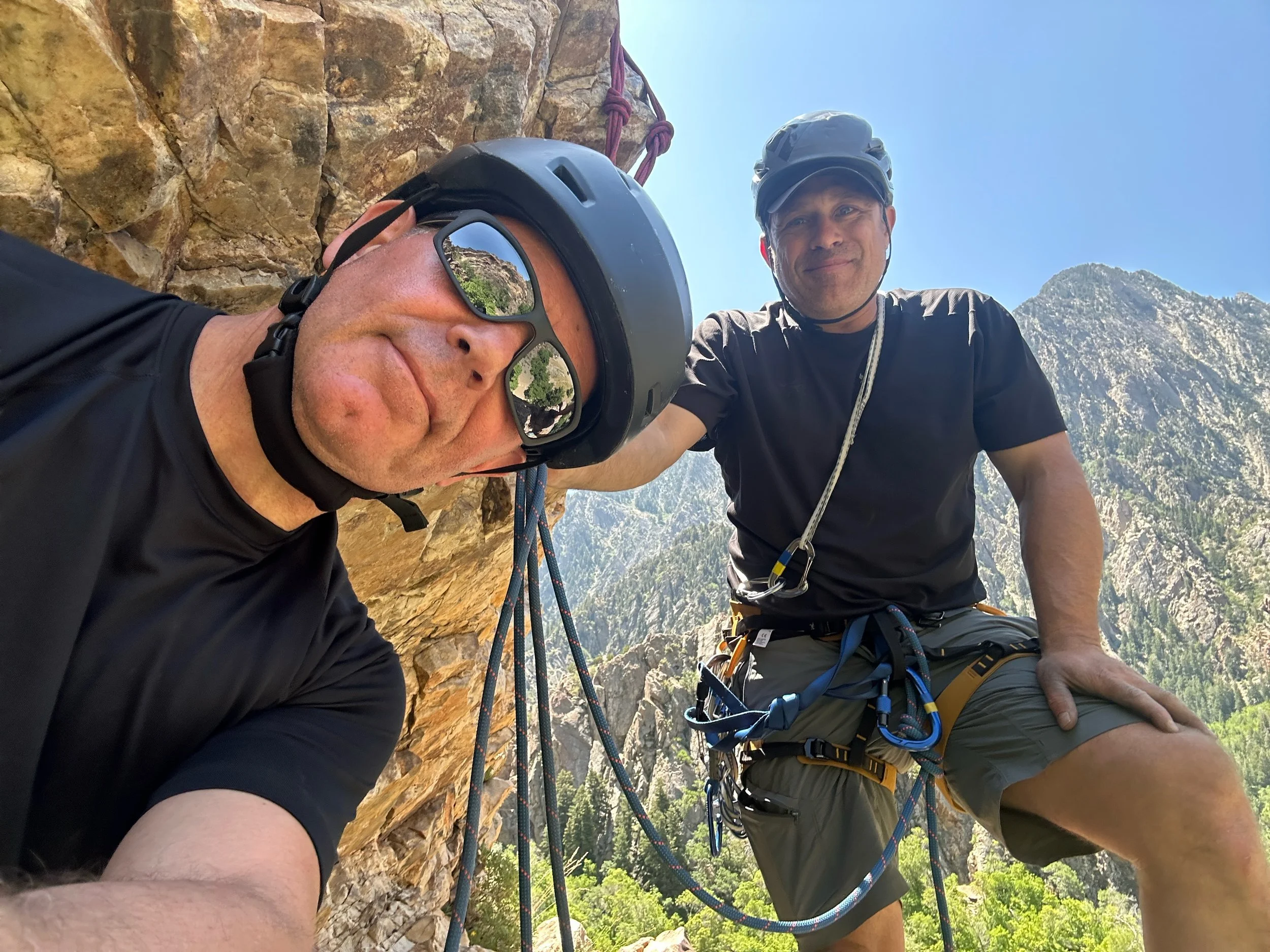 Two rock climbers wearing helmets and harnesses on a cliff, with a mountain landscape in the background; one man is leaning in and wearing sunglasses, while the other is standing and smiling.