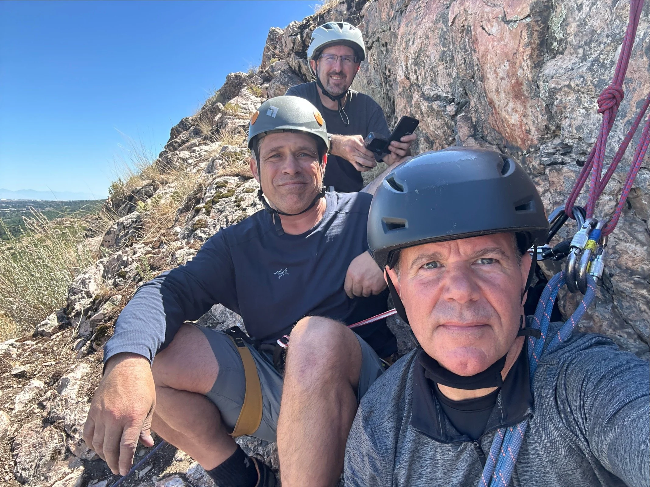 Three men on a rocky mountainside, wearing helmets and climbing gear, taking a selfie with a smartphone, with a clear blue sky in the background.