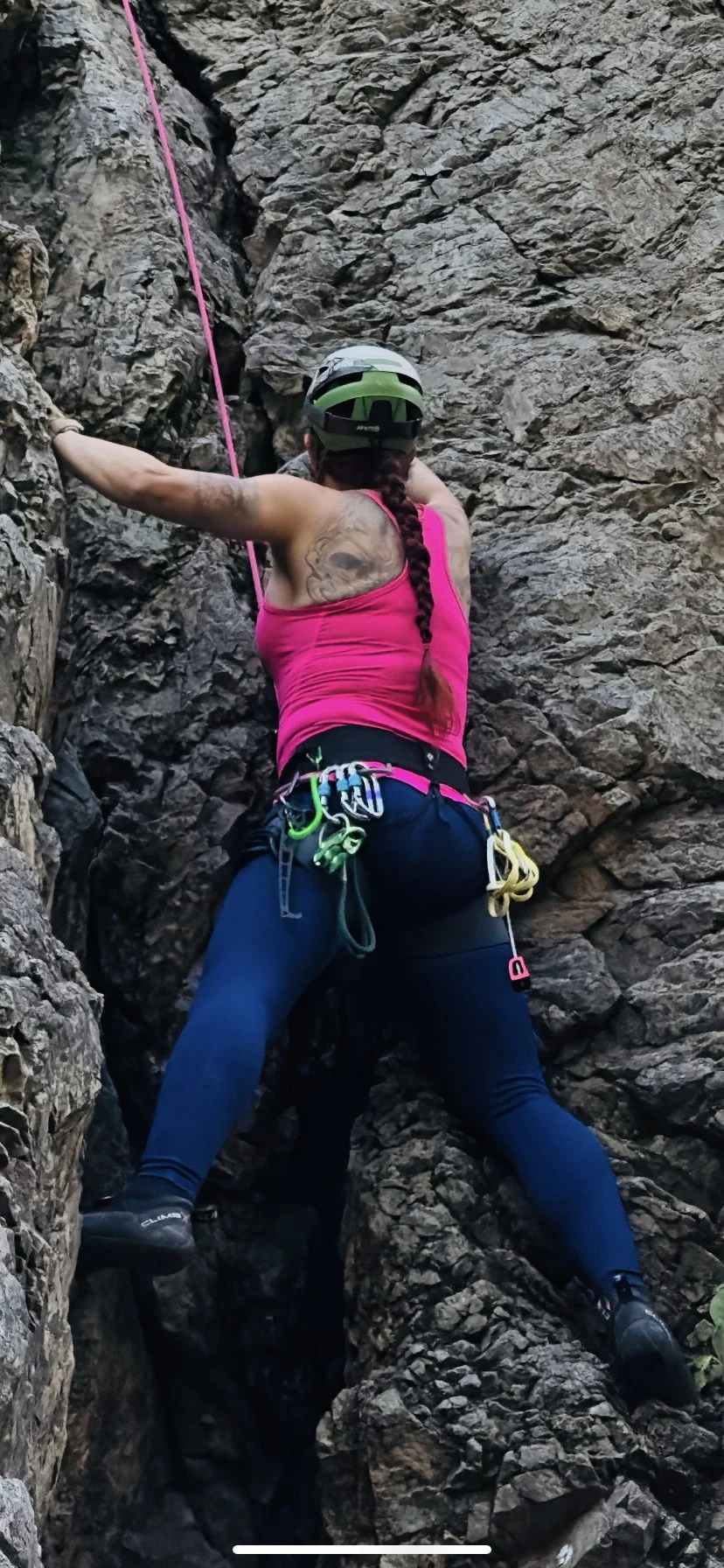 Woman rock climbing on a rugged rock face, wearing a pink tank top, dark pants, climbing harness with carabiners, helmet, and gloves.