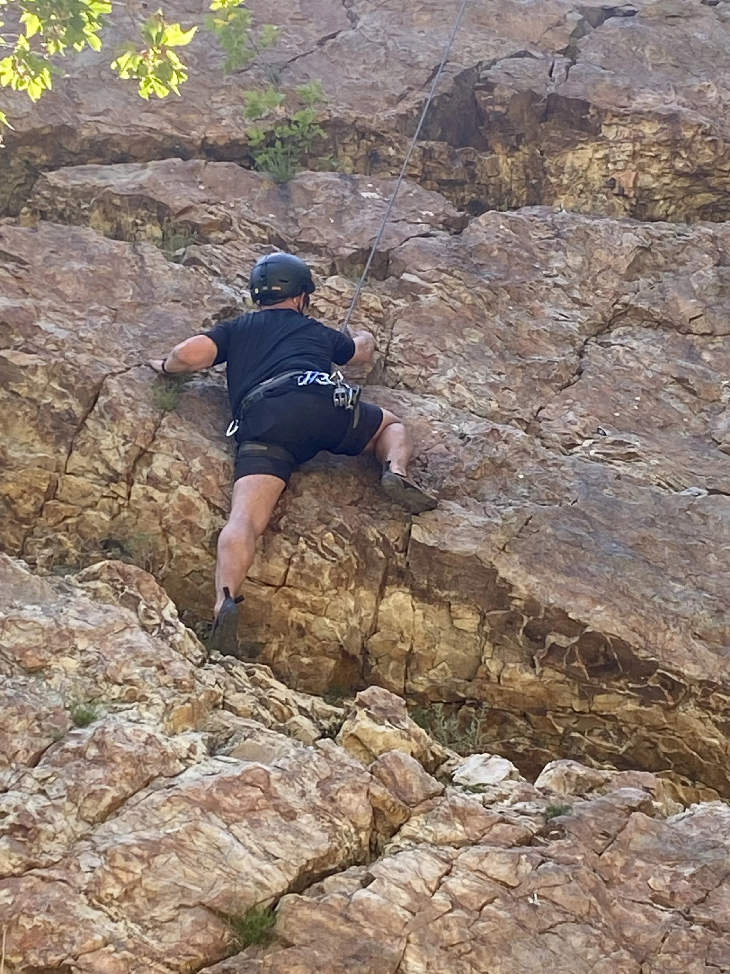 A person wearing a helmet climbing a rocky outdoor wall, holding onto a rope, and wearing a harness attached to the rope.