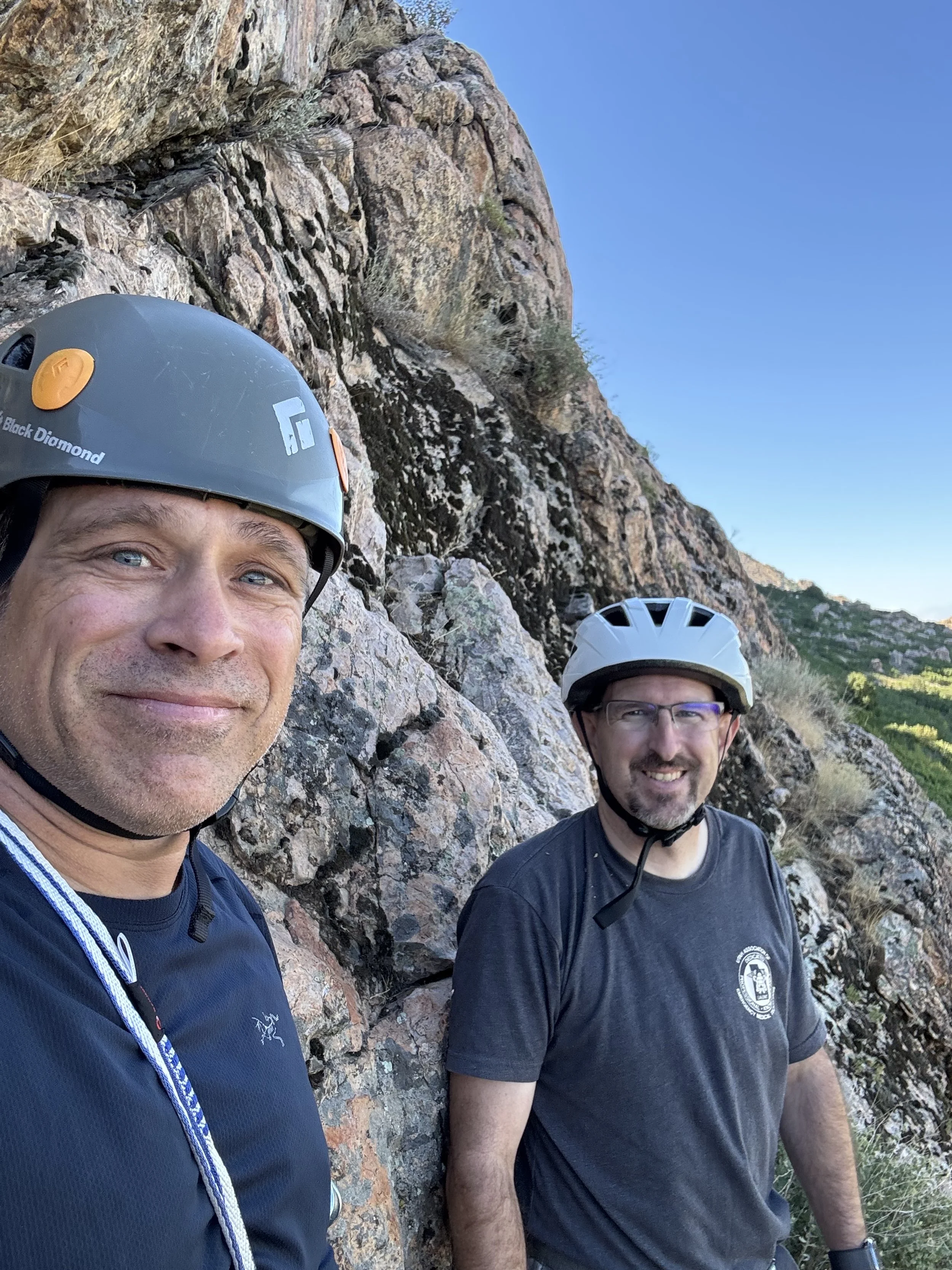 Two men wearing helmets are taking a selfie near a rocky canyon wall with a clear blue sky in the background.