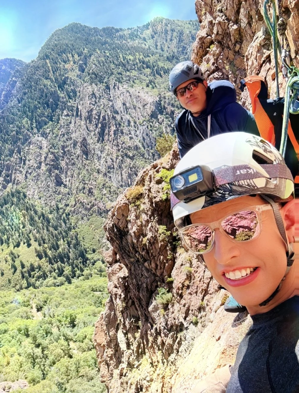 Two climbers on a mountain ridge wearing helmets and sunglasses, with one smiling at the camera, and a lush green valley below with steep rocky cliffs and dense forest in the background.