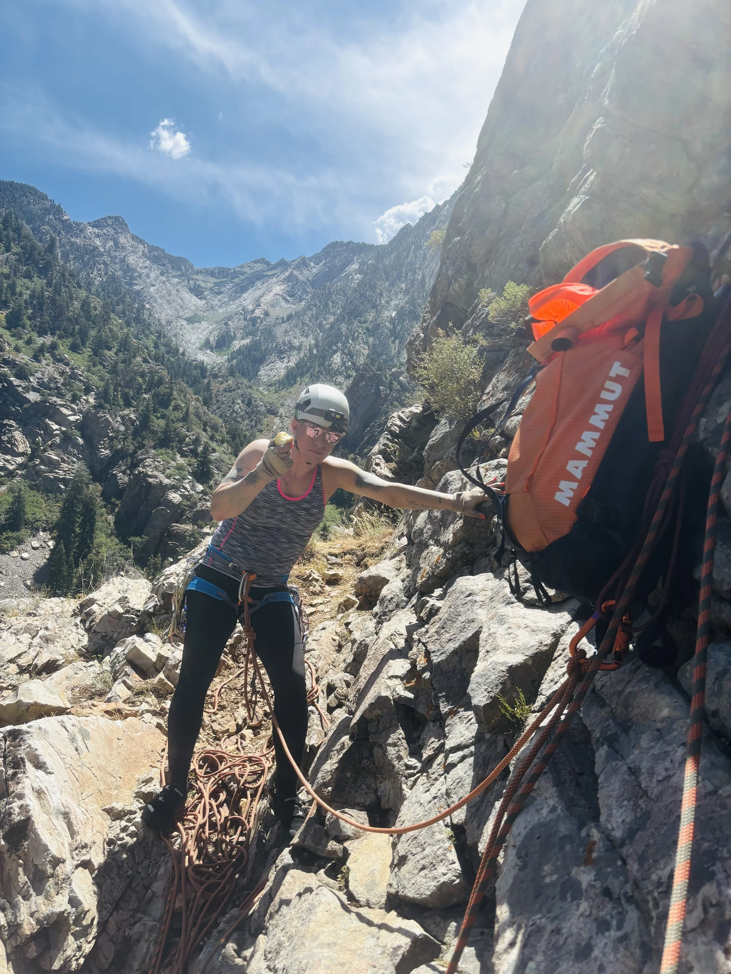A female hiker climbing a rocky mountain trail, wearing a helmet and sunglasses, carrying an orange backpack, with a safety rope, in a mountainous landscape with trees and a bright blue sky.