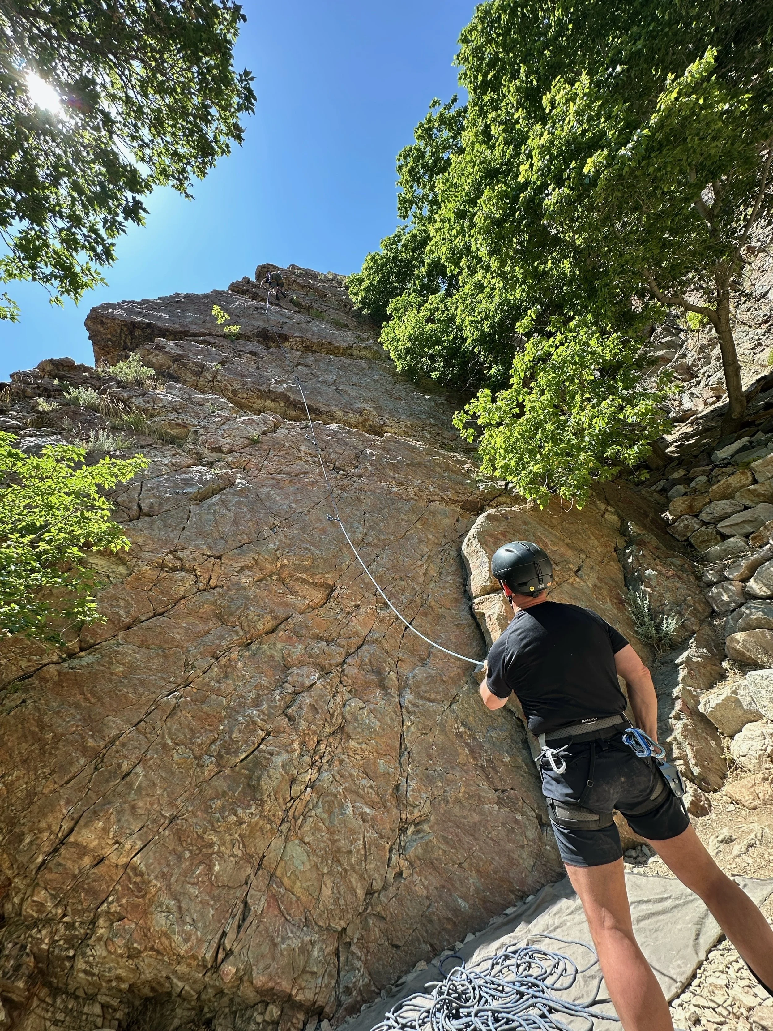 A person wearing a helmet and climbing gear belays a rock climber on a steep outdoor rock face with green trees and a blue sky overhead.