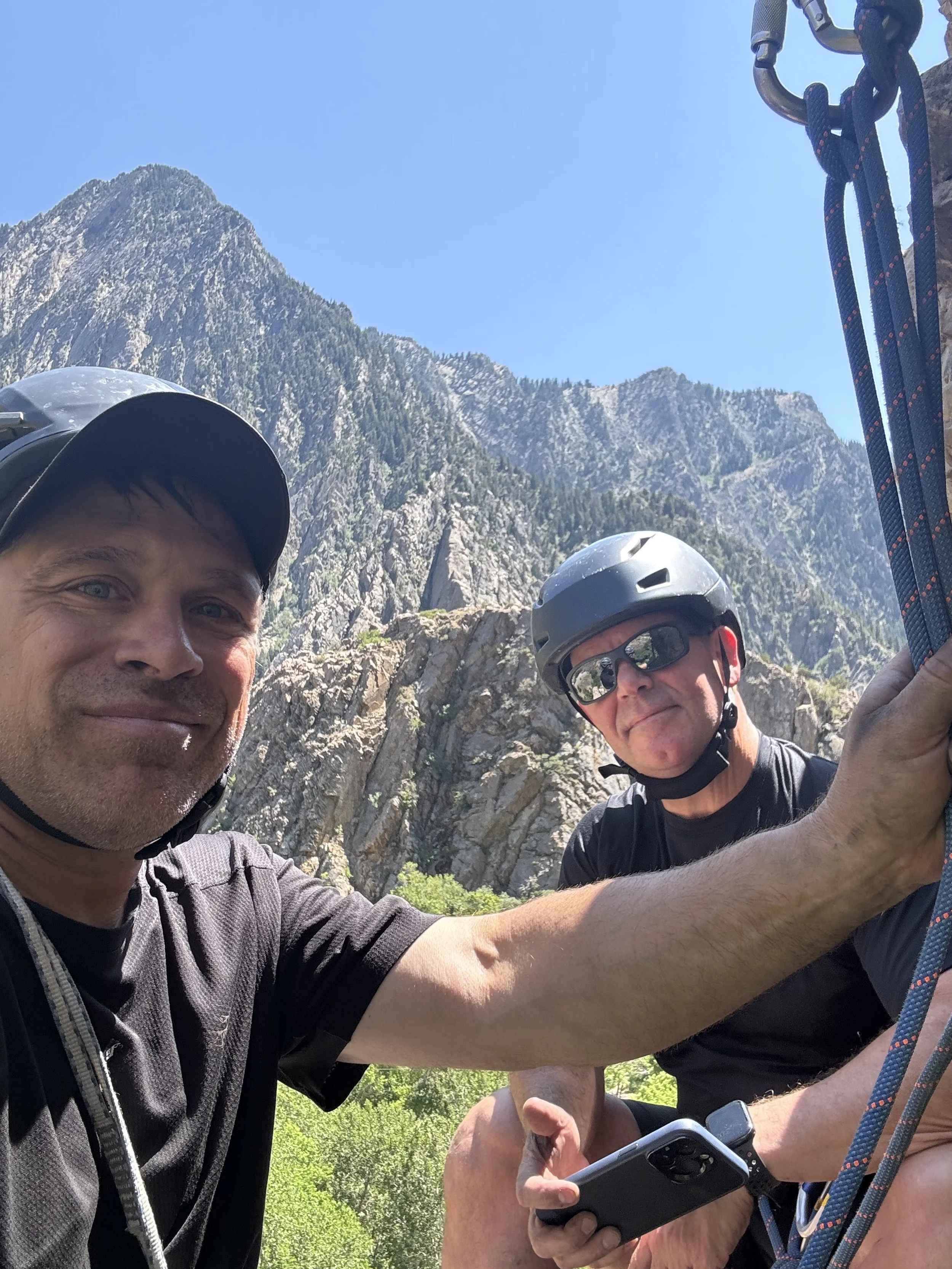 Two men wearing helmets and sunglasses taking a selfie outdoors with mountains in the background.