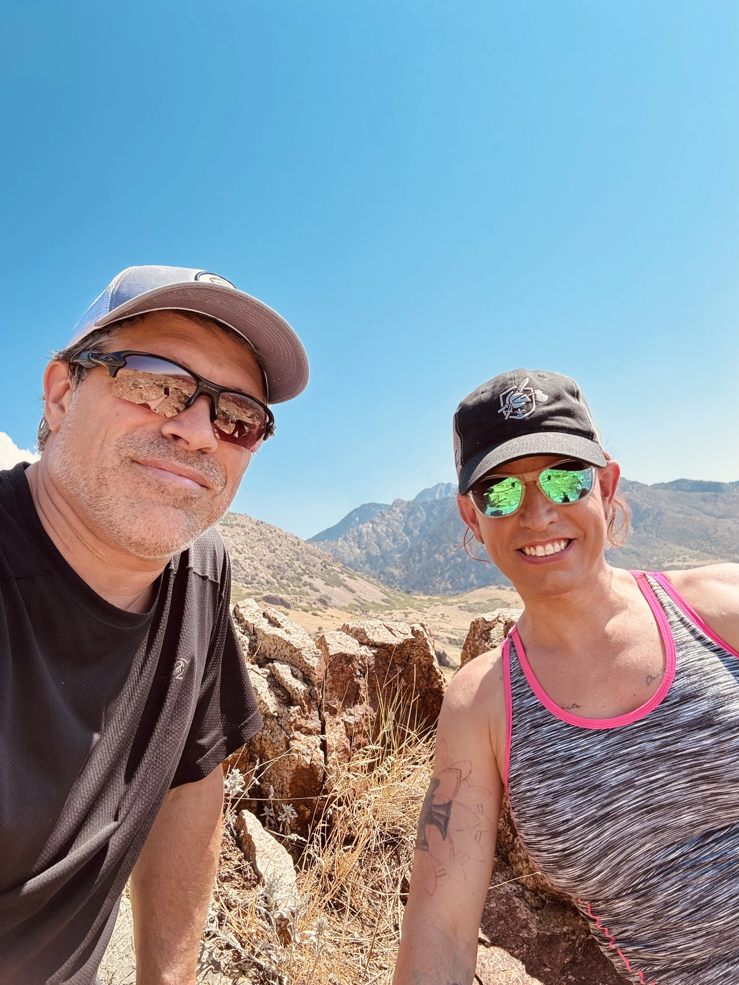Two people, a man and a woman, taking a selfie outdoors in a mountainous desert landscape under a clear blue sky. The man is wearing sunglasses, a cap, and a black shirt, while the woman is wearing mirrored sunglasses, a cap, and a gray athletic top 