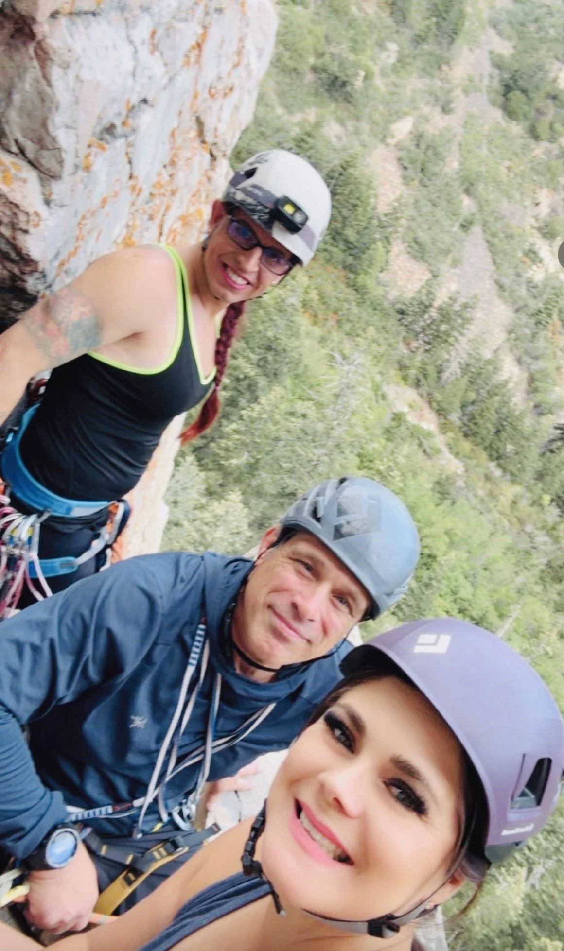 Three climbers wearing helmets and harnesses taking a selfie on a rocky mountain face, with lush green forested landscape in the background.