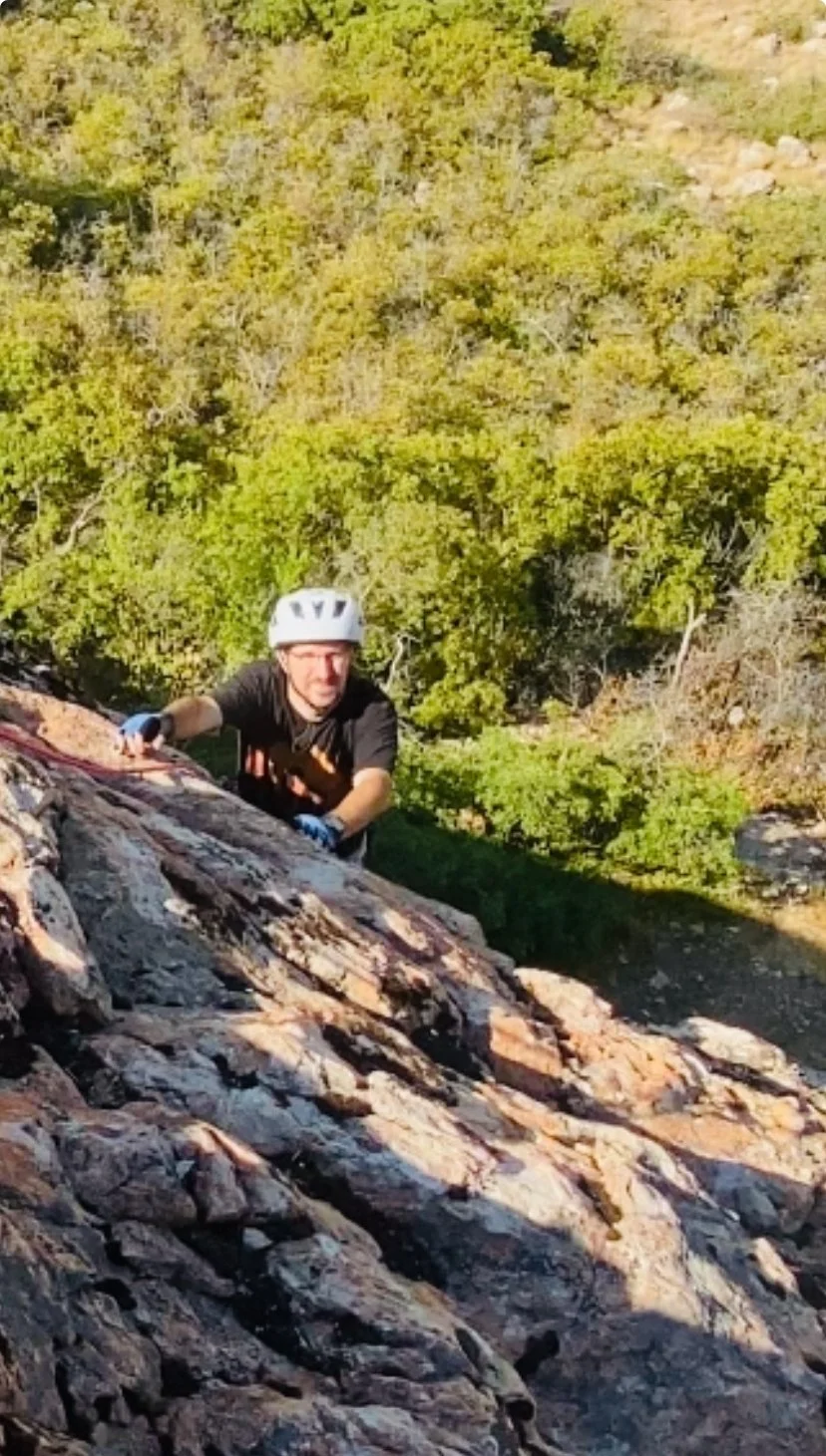 A man wearing a helmet and climbing gear ascending a rocky slope in a natural outdoor setting with trees in the background.